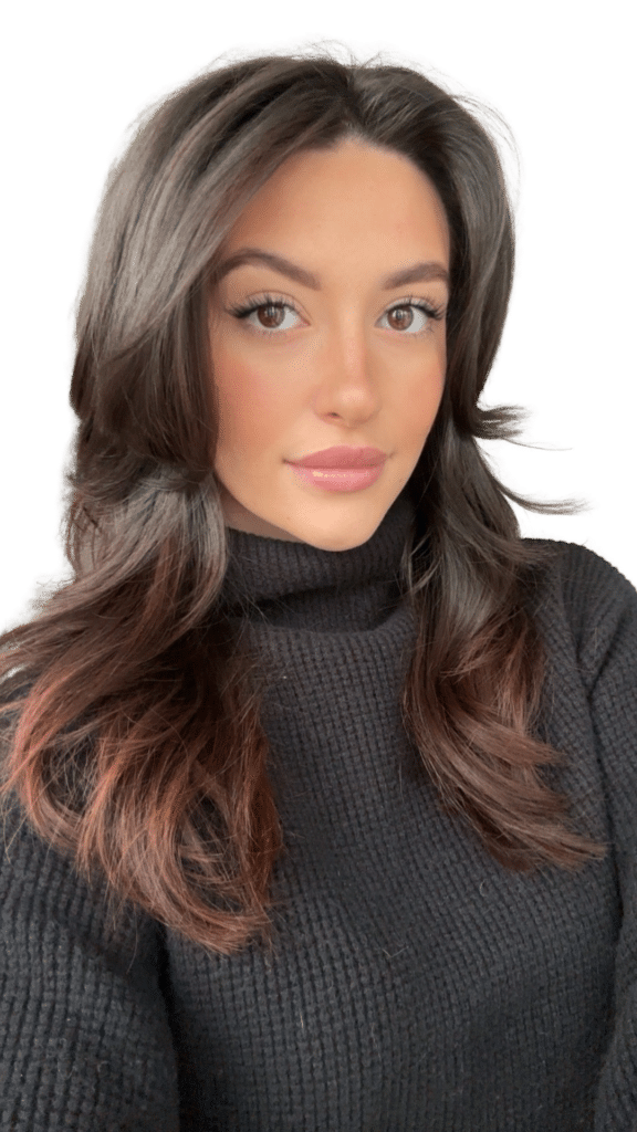 A woman with long, wavy dark hair poses for a selfie. She is wearing a black turtleneck sweater and has a natural makeup look. The background is blurred, highlighting her facial features.