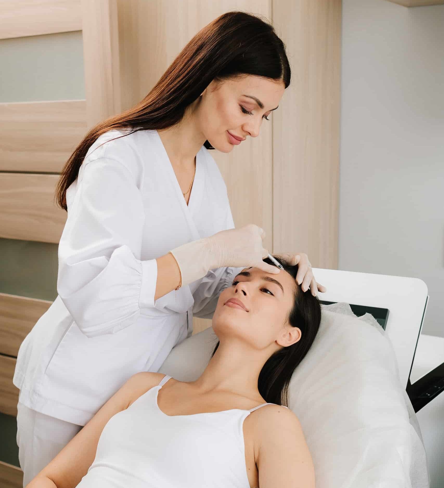 A medical professional in a white uniform is administering treatment to a woman lying on a treatment bed. The setting appears to be a modern clinic, with neutral-colored walls and equipment.