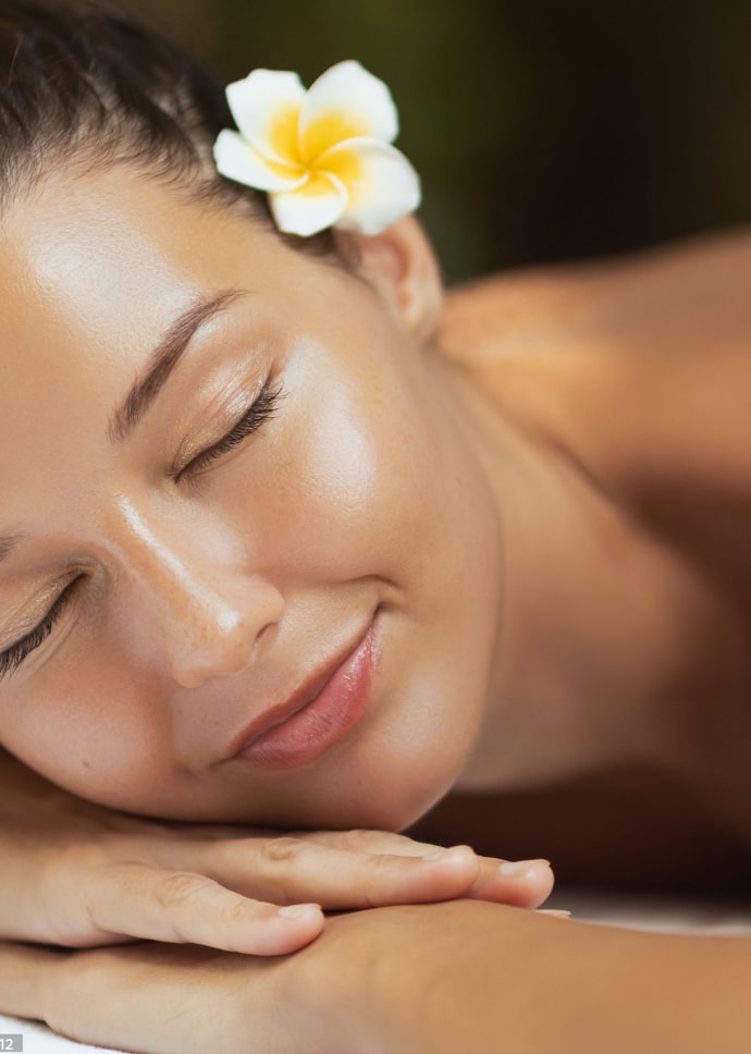 A woman with long brown hair and a white flower in her hair is resting peacefully while lying on a table. She has a relaxed expression with her eyes closed, showcasing smooth, glowing skin.