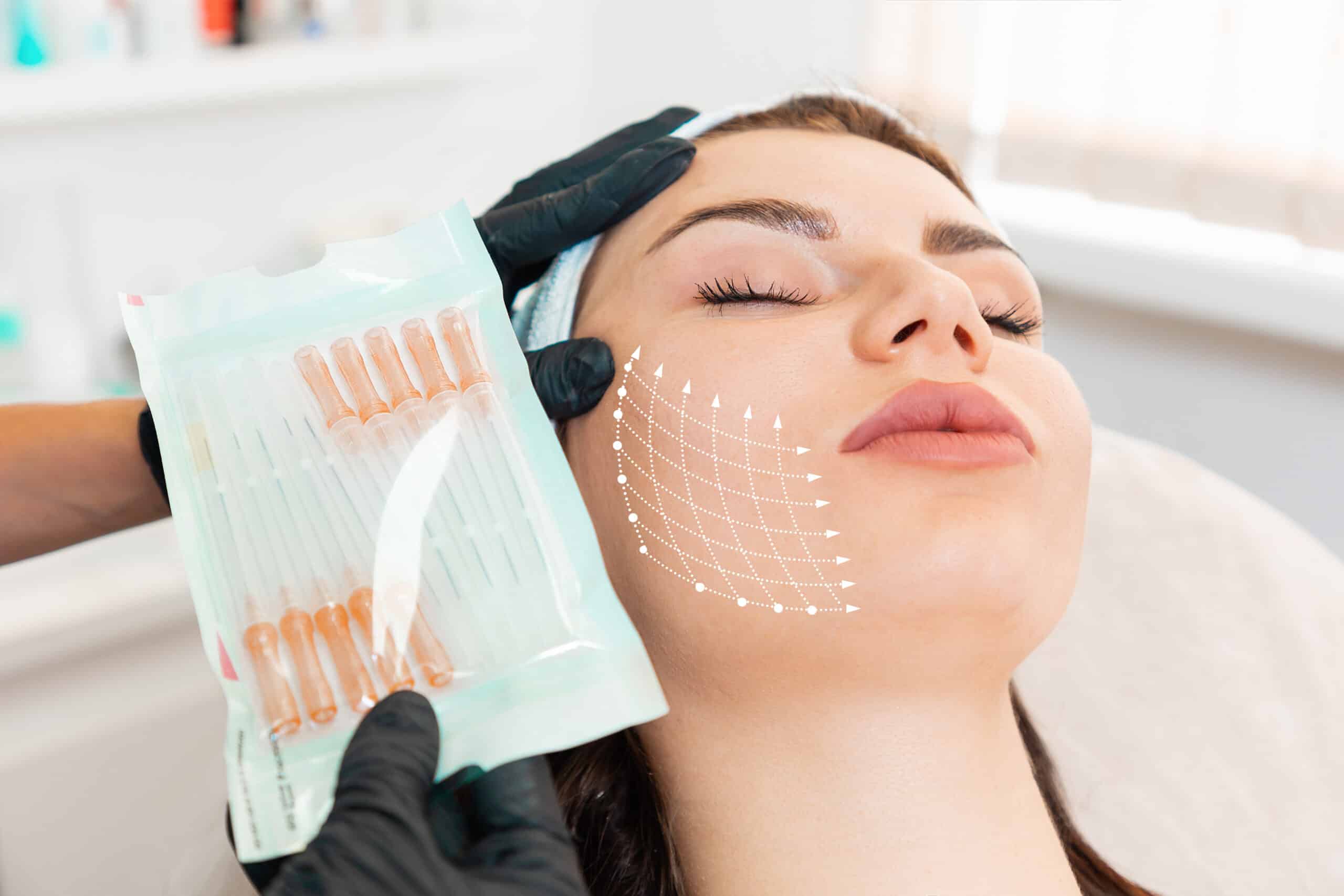 A woman receiving a facial treatment while a technician holds a pack of cosmetic serum vials next to her face, which has overlay markings for skin application. The setting is a modern skincare clinic.