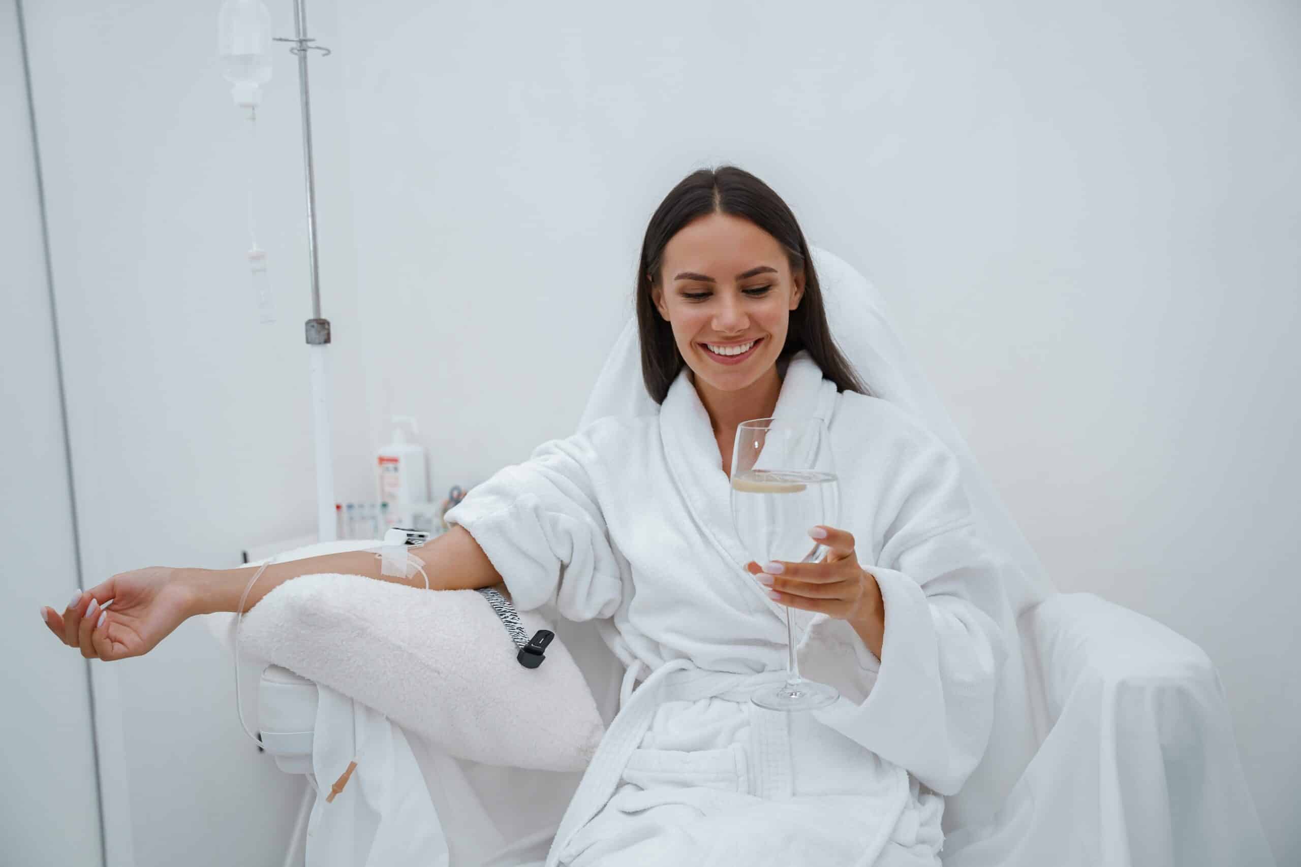A woman wearing a white robe sits comfortably in a treatment chair, smiling while holding a glass of drink. An IV drip is connected to her arm, and the background is minimalist and bright with white walls.