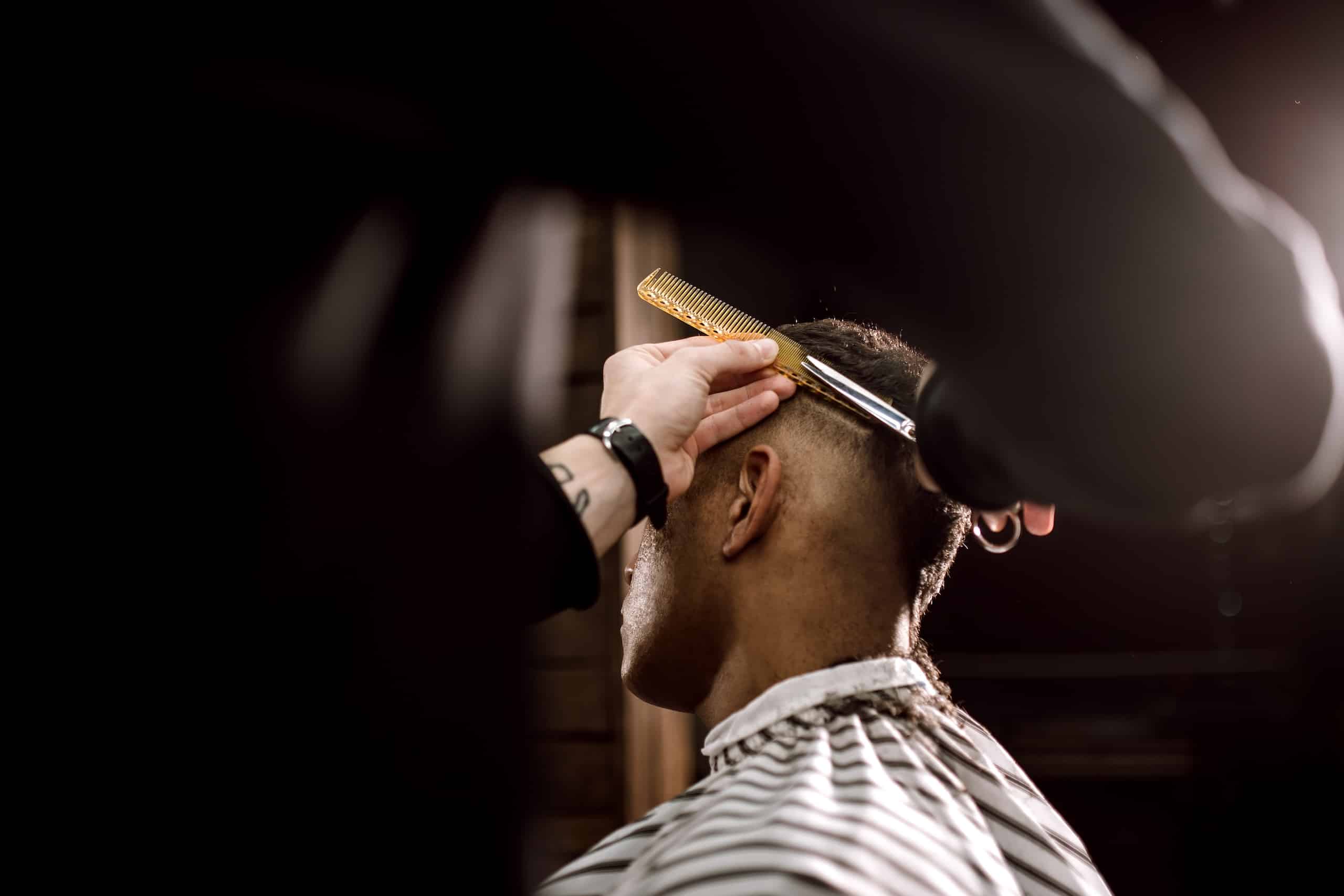 A barber using a comb and scissors to style the hair of a client with a fade haircut, viewed from behind. The client wears a striped cape and is seated in a dimly lit barbershop.