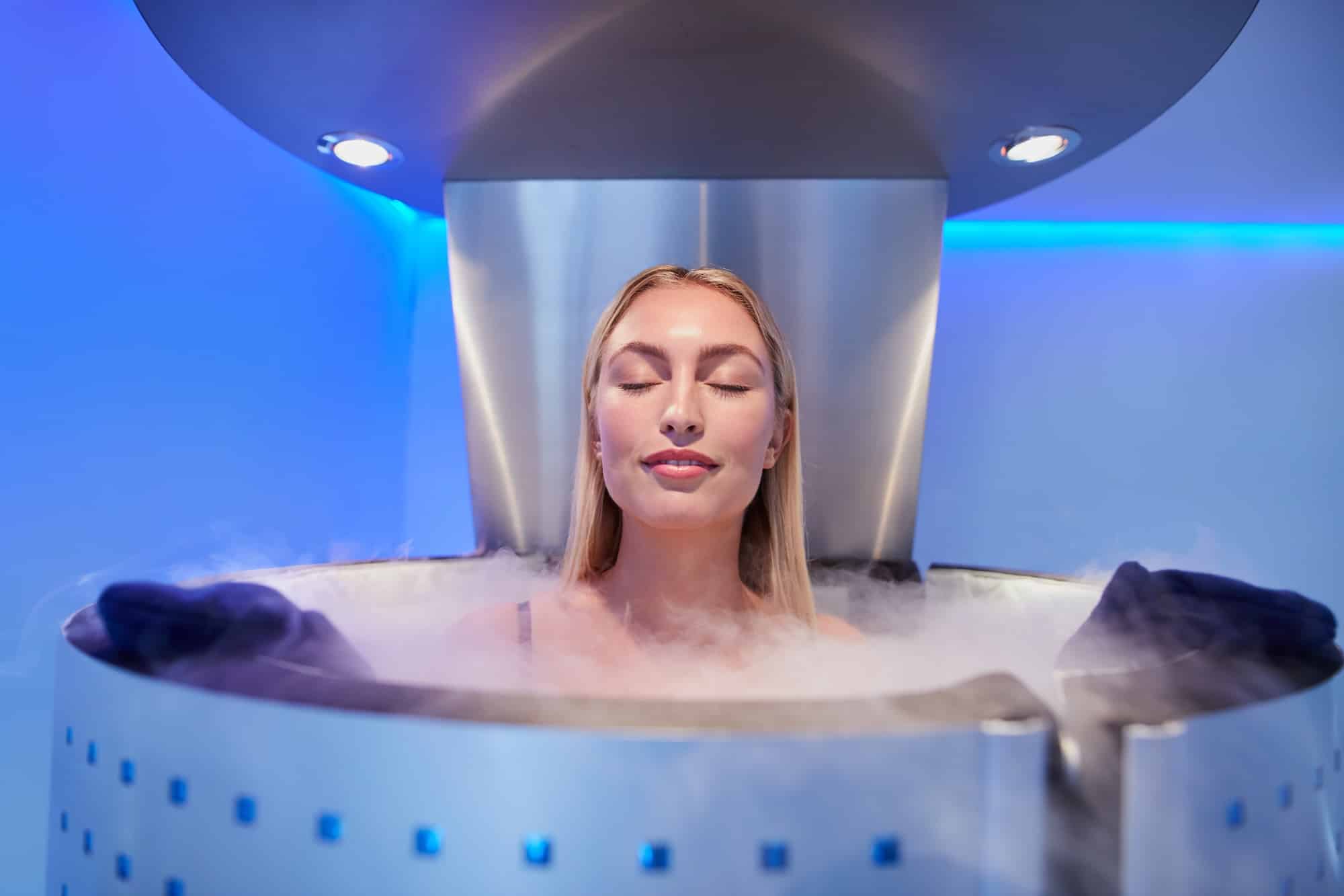 A woman with blonde hair relaxing in a cryotherapy chamber, surrounded by fog. The chamber features sleek metallic design and is illuminated by blue lights.