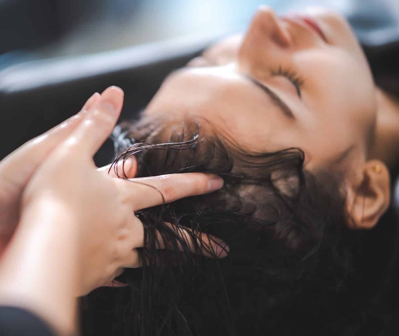 A close-up of a woman receiving a hair treatment, with a person's hand gently massaging her scalp. The woman appears relaxed, with her eyes closed. The background is softly blurred, emphasizing the intimate salon setting.