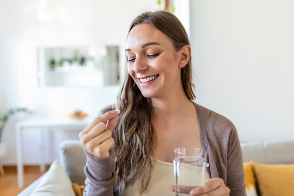 A woman sitting on a couch holds a clear glass of water in her right hand and examines a small tablet in her left hand. She has long brown hair and a slight smile, with a softly decorated living space in the background.