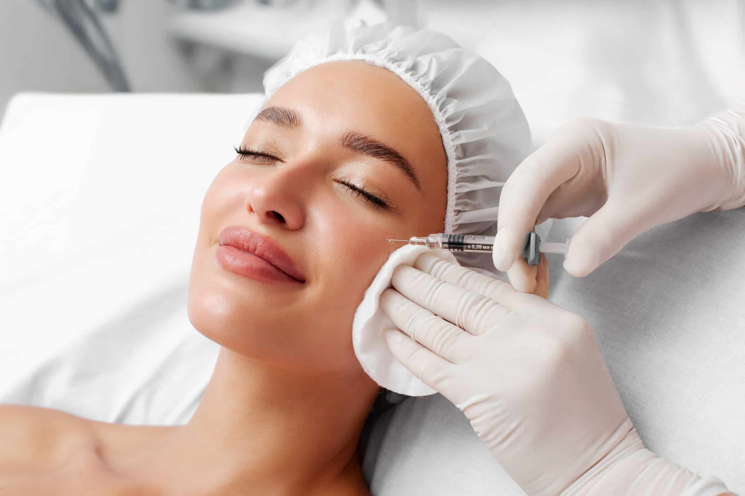 A woman lying on a treatment table with her eyes closed, smiling. A medical professional, wearing gloves, is applying a cosmetic injectable to her cheek while holding a cotton pad. The setting appears clinical and well-lit.
