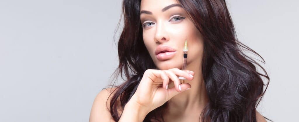 A woman with long, dark hair holds a syringe near her face, lightly touching her cheek. She has a neutral expression and visible make-up, set against a plain light background.