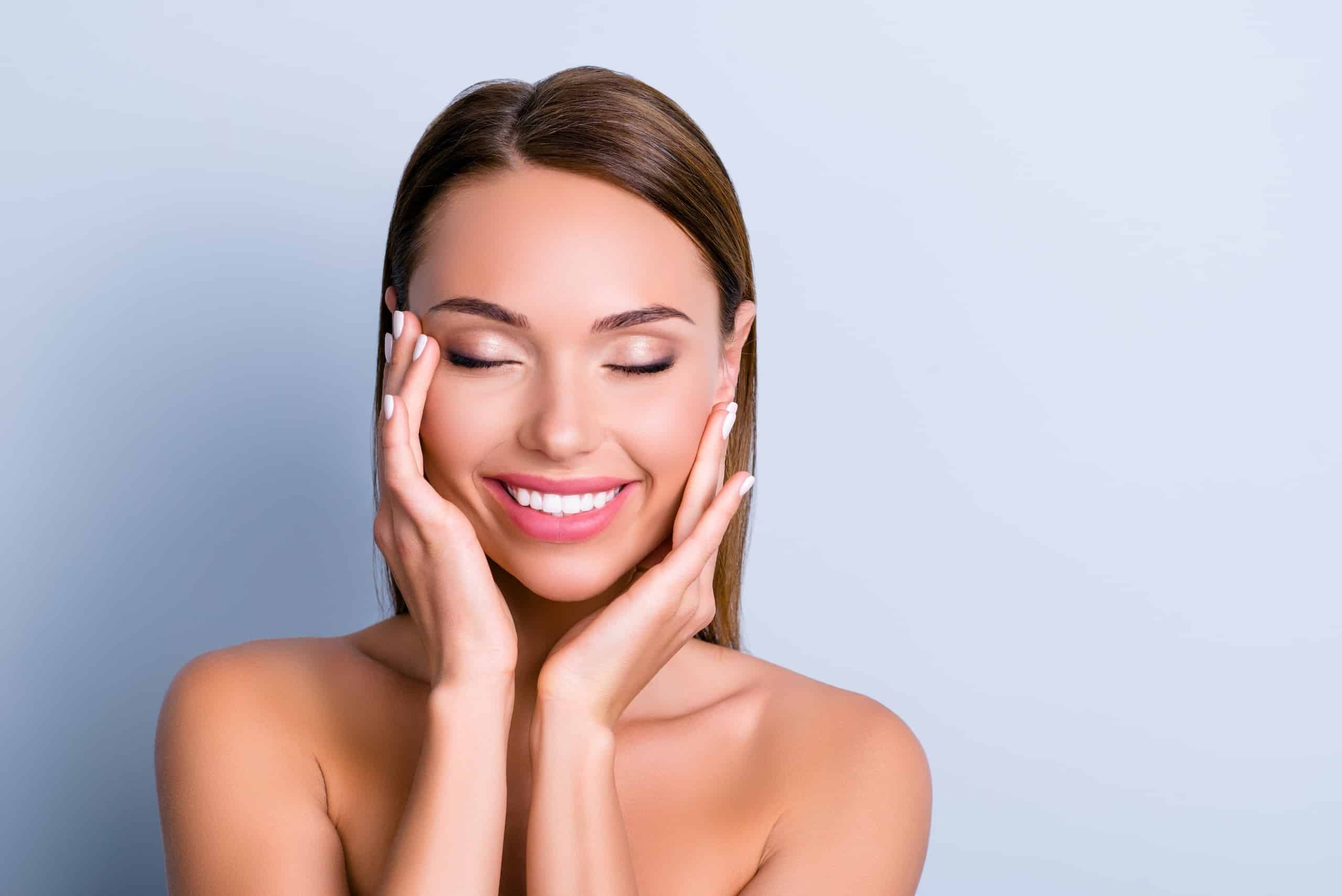 A woman with long brown hair is smiling and has her eyes closed while gently holding her face with both hands. She is posed against a light blue background and has a natural, clean look, with no visible makeup, highlighting her clear skin.