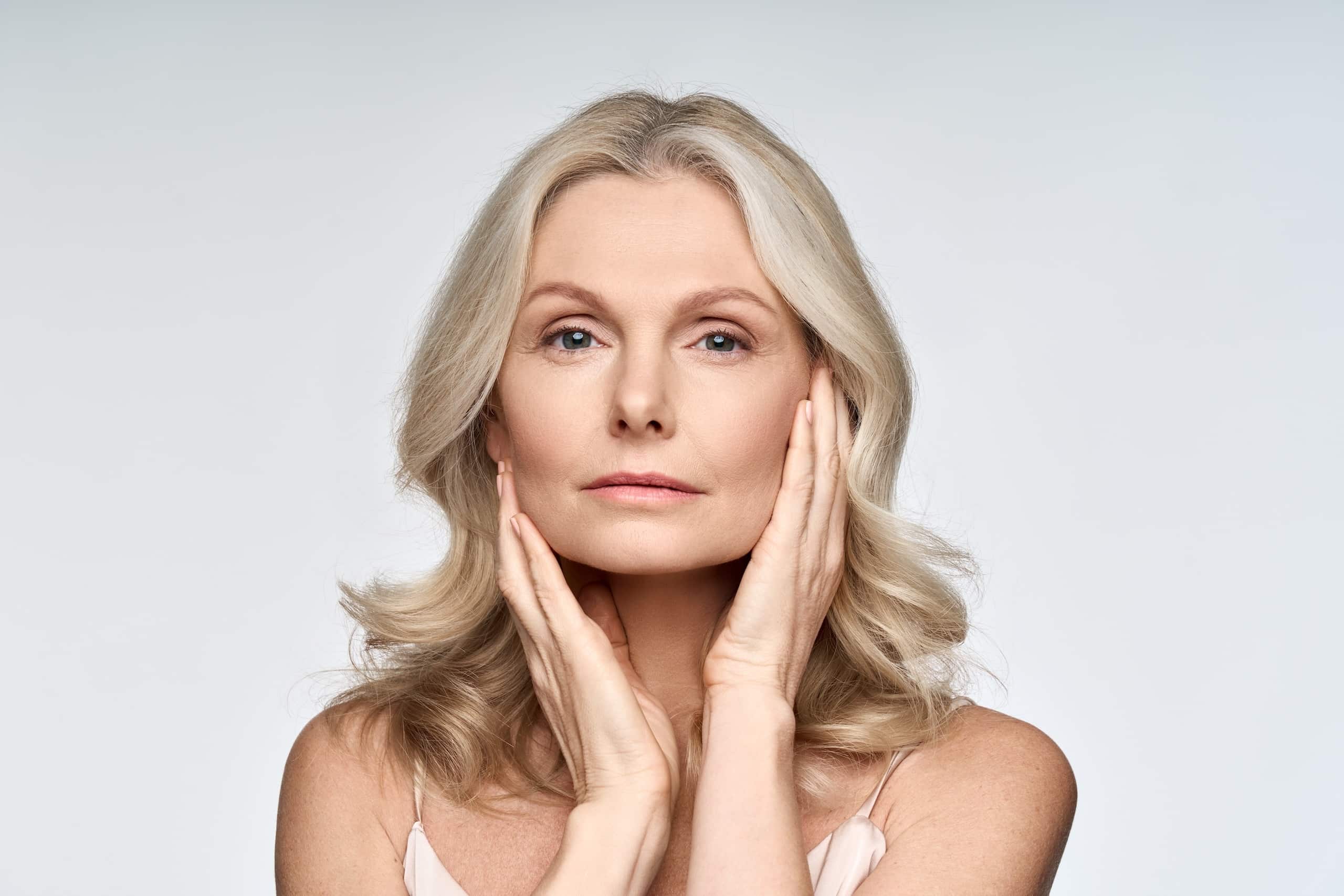 A woman with long, light blonde hair poses with her hands on her face, gently holding her cheeks. She has a neutral expression and is wearing minimal makeup. The background is a soft gray, creating a clean and simple aesthetic.