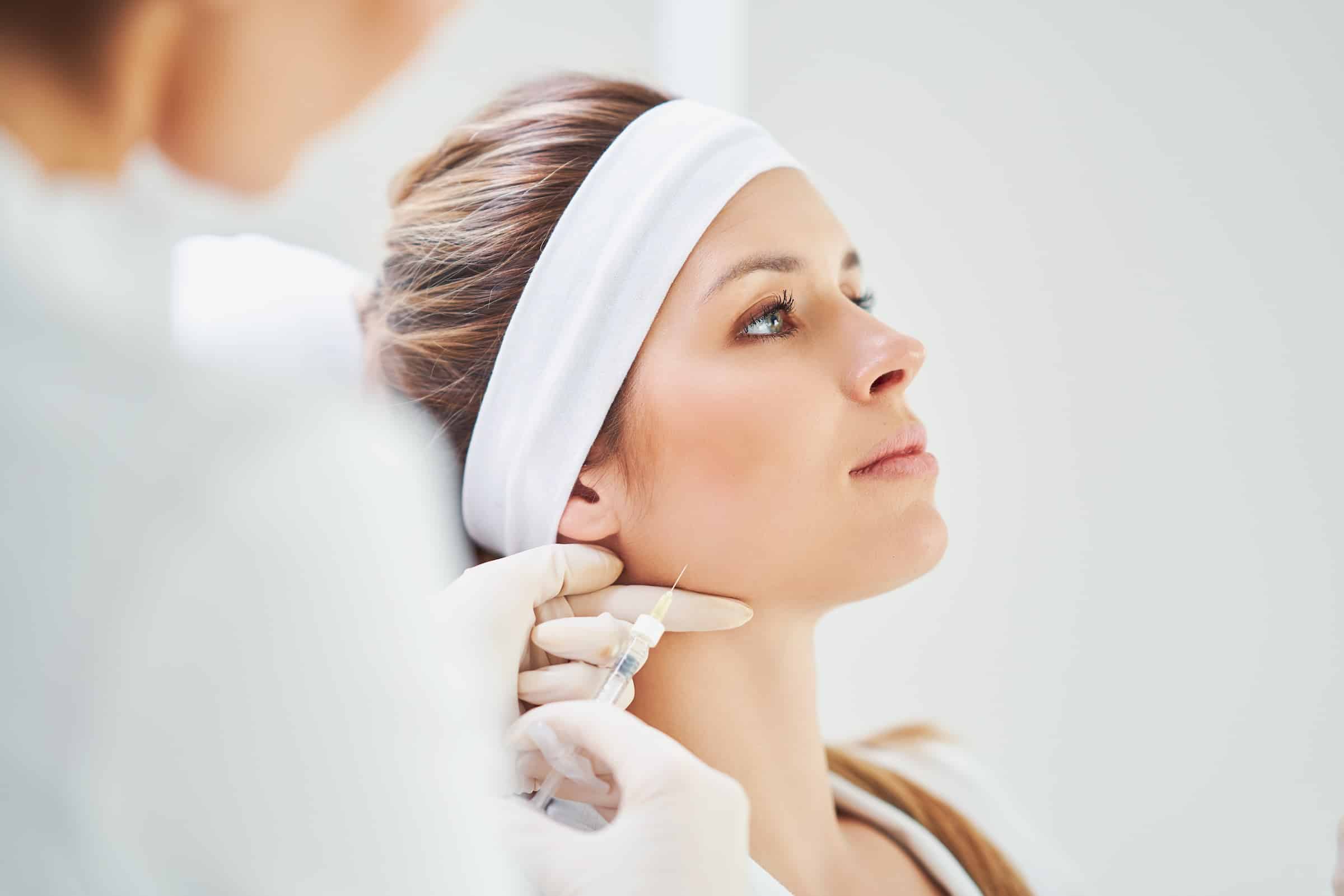 A woman receiving a cosmetic injection in a skincare clinic. She is seated with a headband on, looking calmly to the side as a professional administers the treatment.