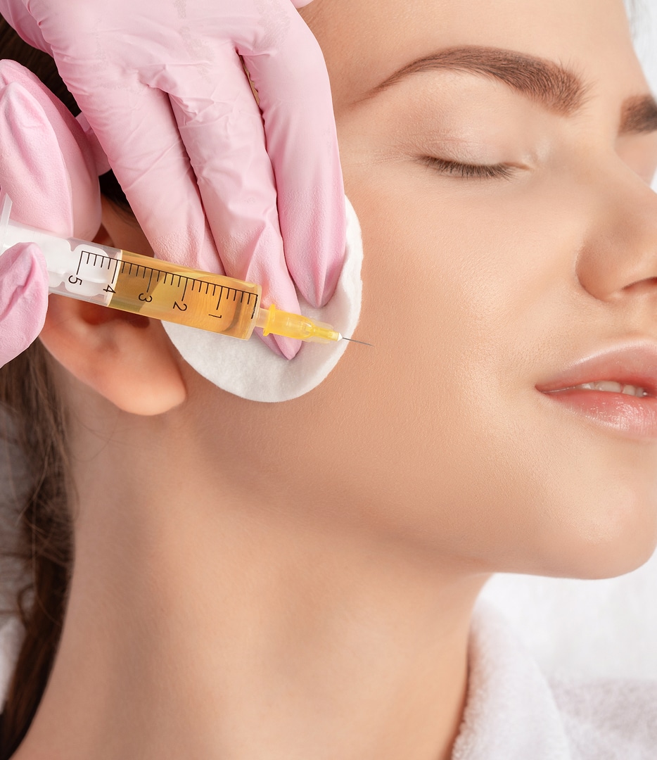 A professional wearing pink gloves holds a syringe near a woman's cheek, who has her eyes closed, as part of a facial treatment in a clinical setting.