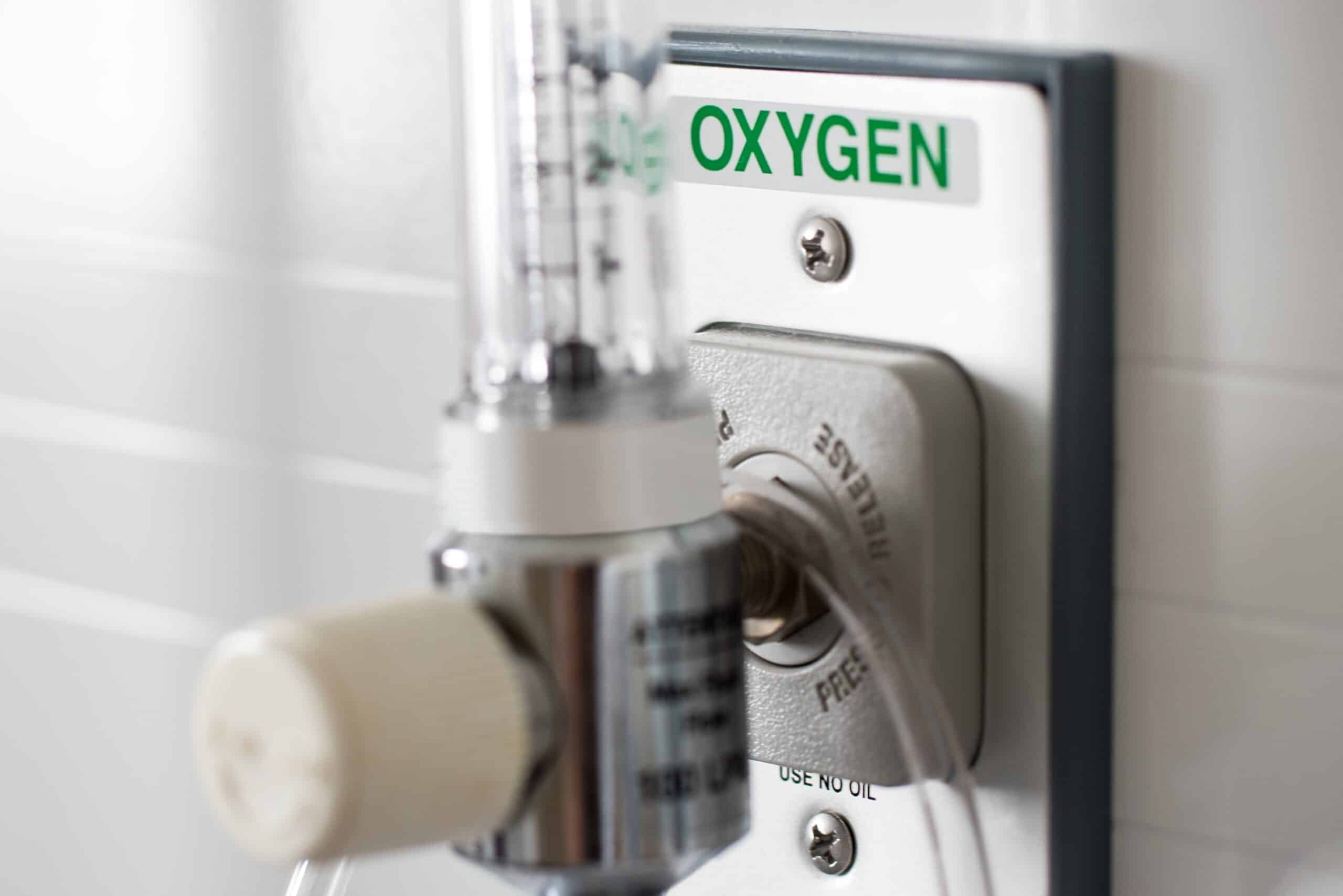 A close-up view of an oxygen supply unit mounted on a wall. The unit features a clear flowmeter and a knob for adjusting the flow. The white wall panel is labeled "OXYGEN" in green letters, indicating the oxygen source.