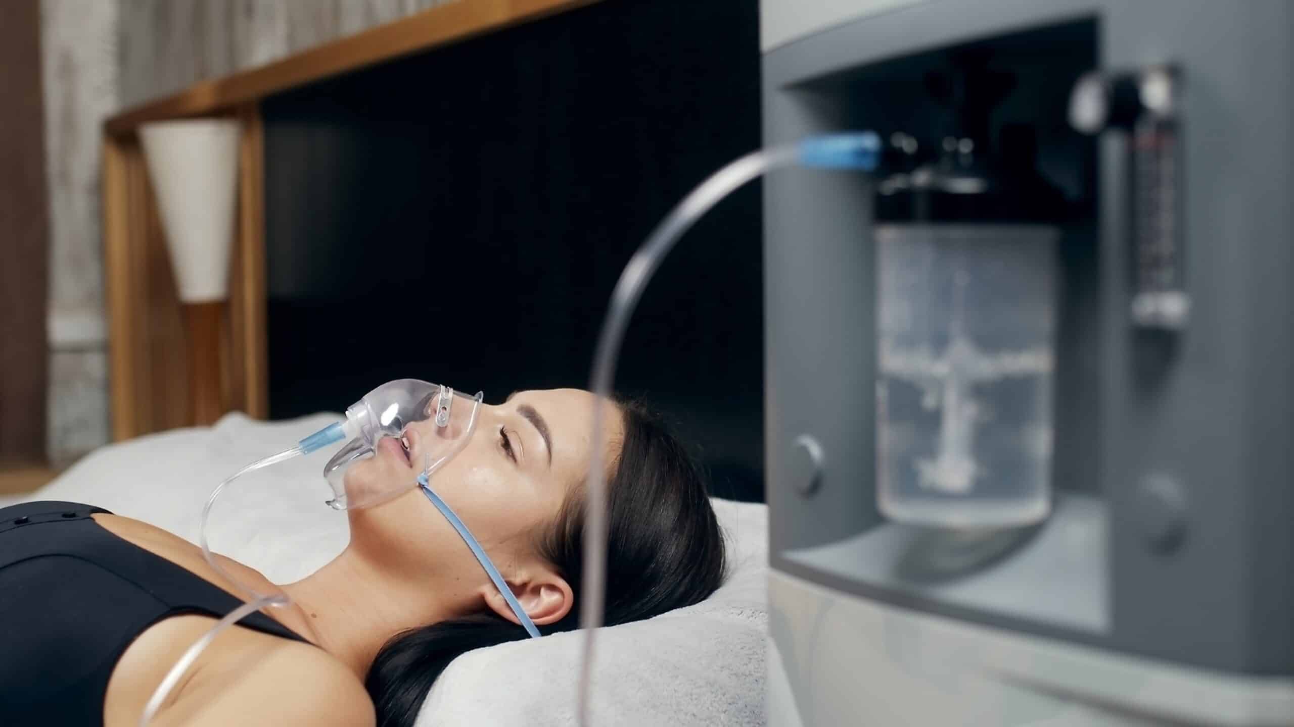 A woman lying on a bed with a face mask connected to an oxygen machine, which is situated beside her. The room has a modern, neutral decor with wooden accents, and the woman appears relaxed with her eyes closed.