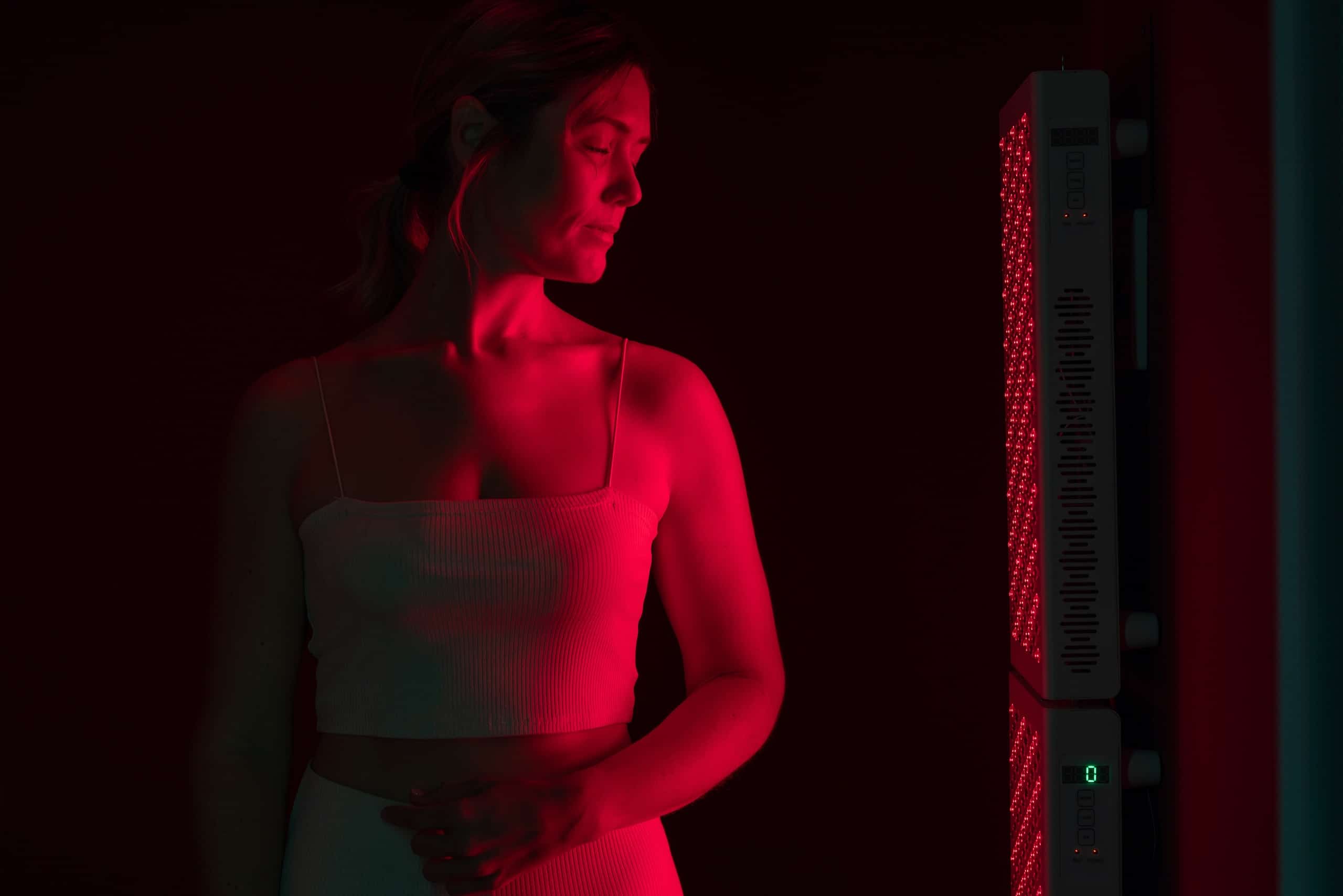 A woman wearing a white tank top stands near a vertical device emitting red light in a dim environment. She gazes thoughtfully at the device, which has a grid pattern illuminated. The overall lighting creates a warm, red hue around her.