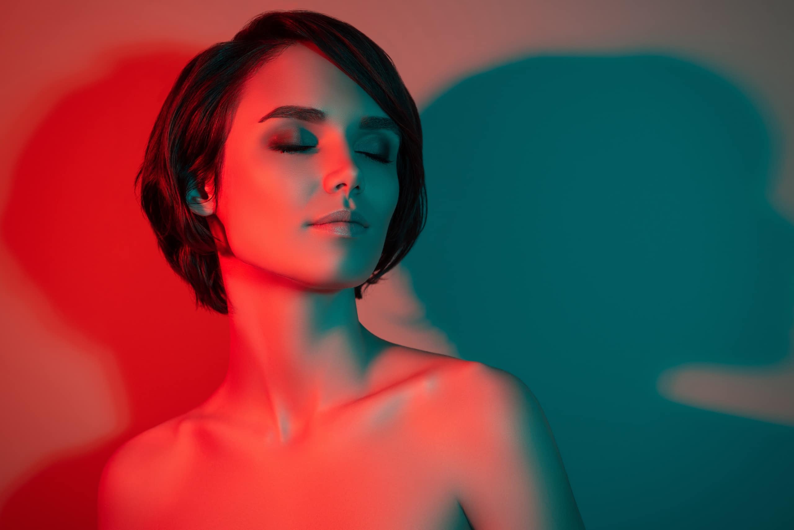 A woman with short, dark hair is depicted against a backdrop of red and blue lighting. Her eyes are closed, and she has a calm expression. The lighting creates contrasting shadows on her face and shoulders, emphasizing her features.