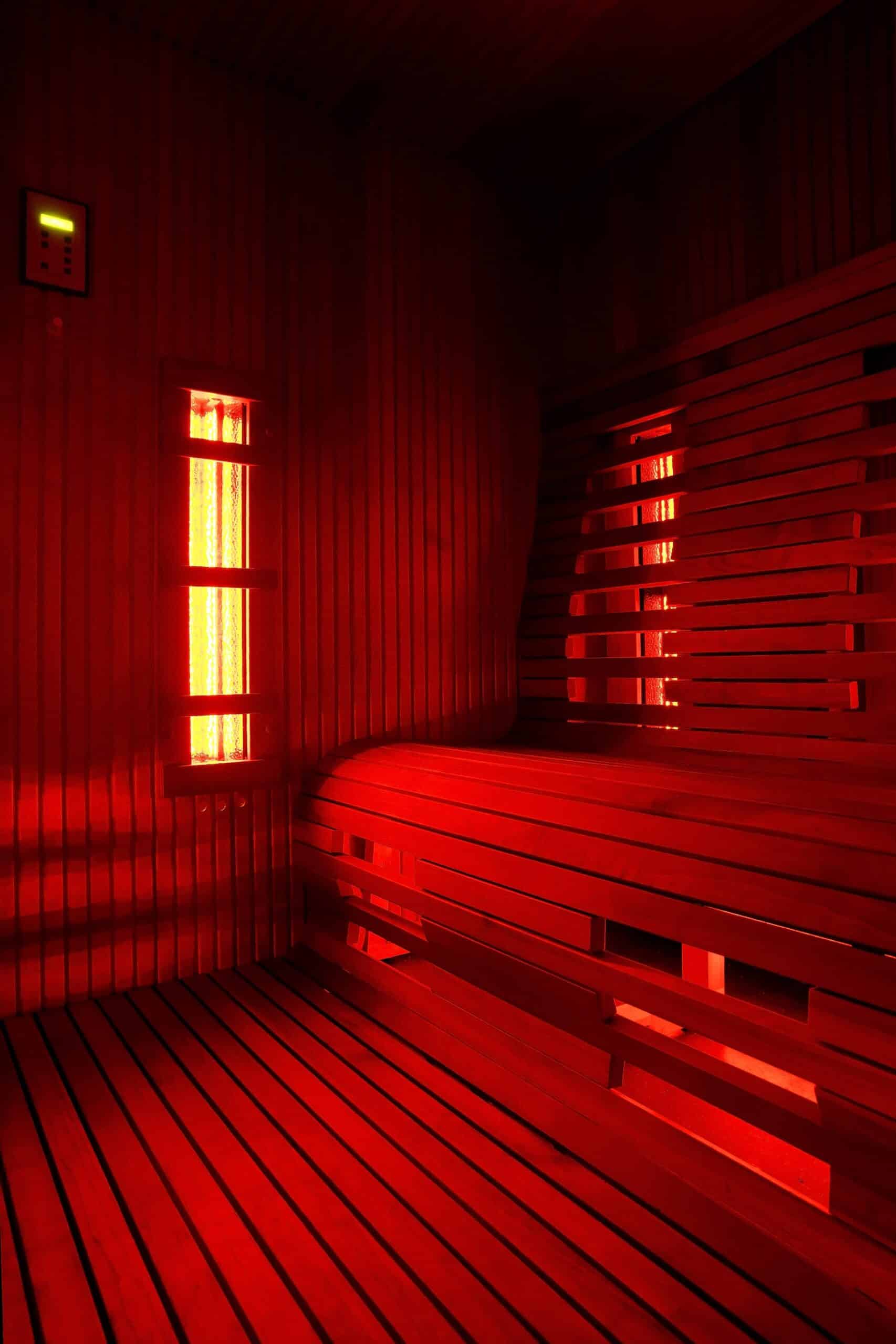 Interior of a sauna featuring wooden benches and wall paneling, illuminated with red light. Two narrow windows allow light to enter, highlighting the sauna's wooden structure and creating a warm atmosphere.