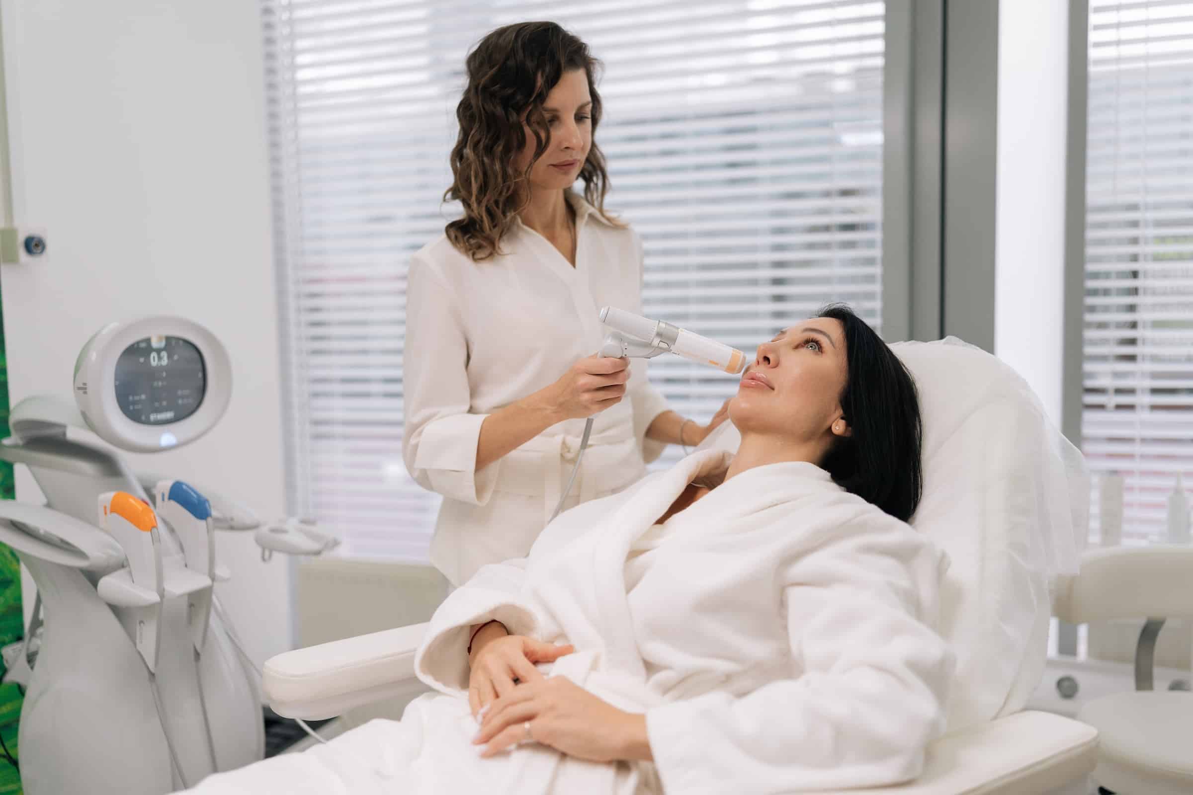 A woman in a white robe receives a skincare treatment while lying on a treatment bed. Another woman, wearing a white outfit, uses a handheld device on her face in a modern clinic setting.