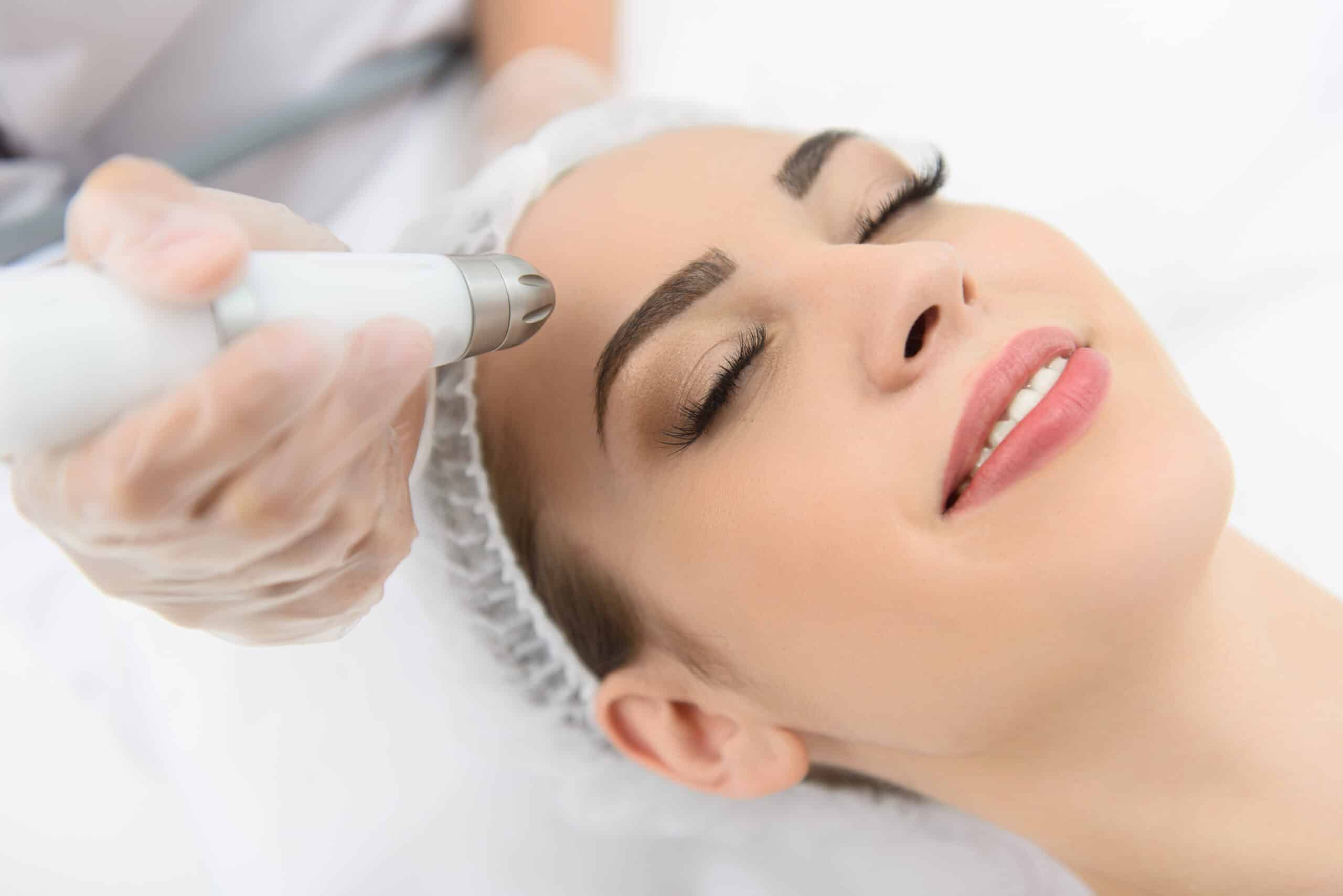 A woman receiving a skincare treatment while lying down. A technician in gloves uses a handheld device near her forehead. The woman has her eyes closed and is smiling slightly. She is wearing a hairnet, and the background is softly blurred.