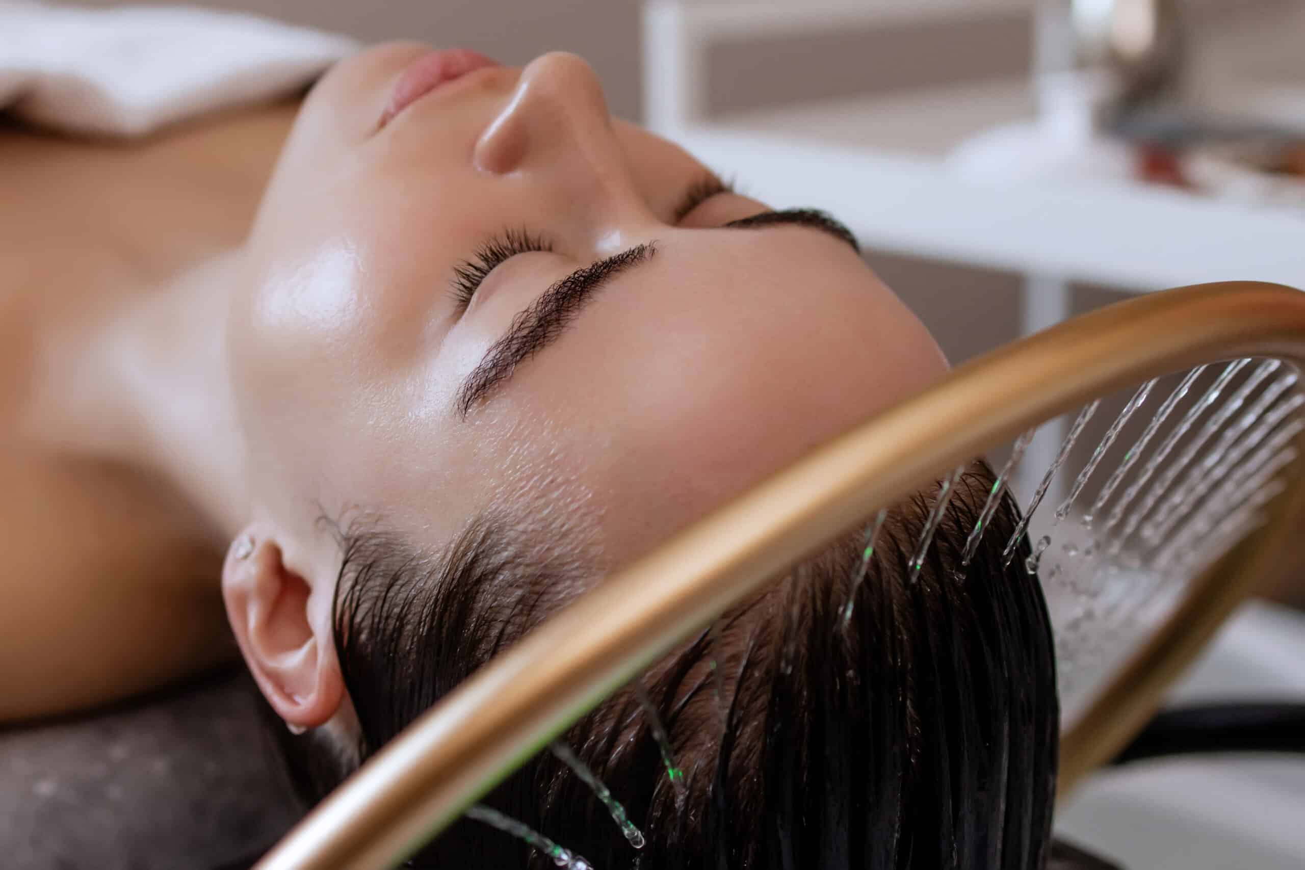 A woman relaxes in a salon chair with her eyes closed, undergoing a hair treatment. Water is running through her hair, and she is positioned under a salon dryer with a gold rim. The background features a clean, modern salon environment.