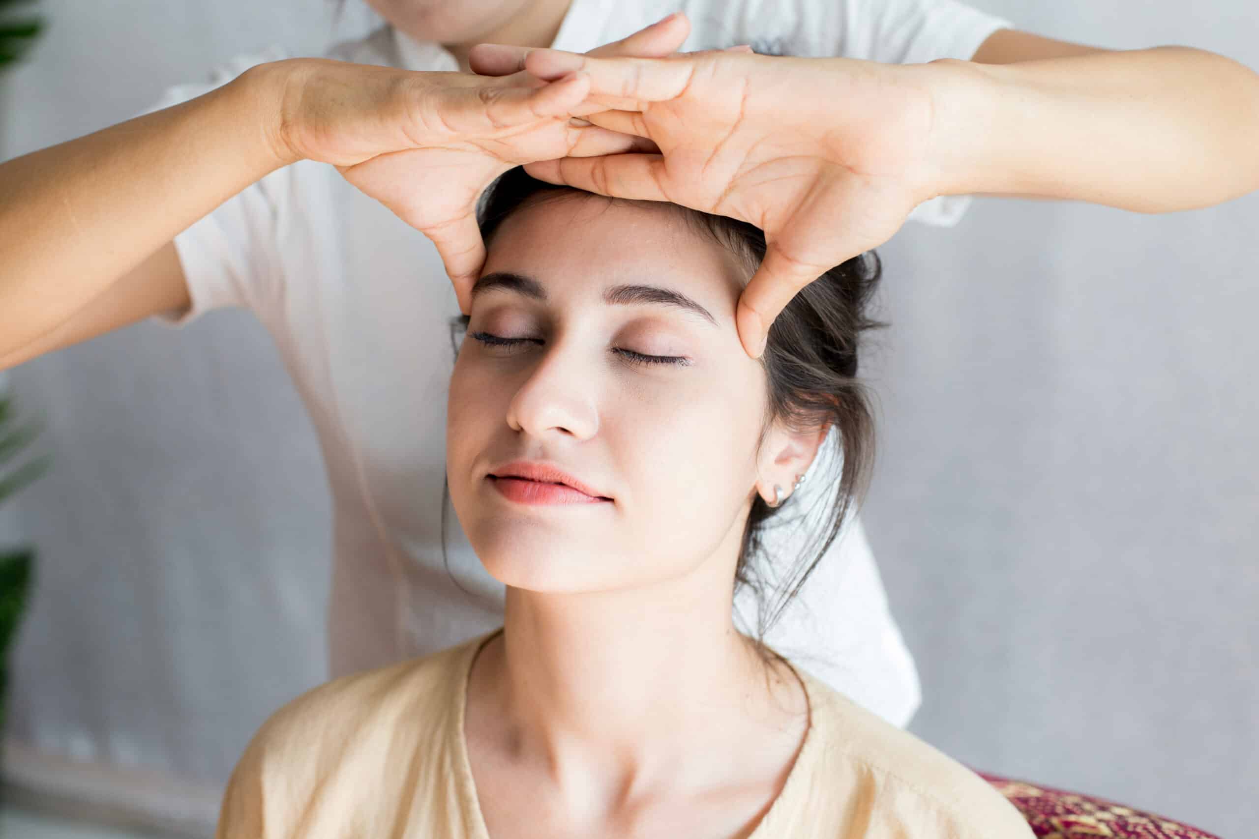 A woman receiving a head massage while sitting comfortably. A therapist's hands are positioned on the woman's forehead, and she has her eyes closed, suggesting relaxation. The setting appears calm with soft lighting and minimal background.