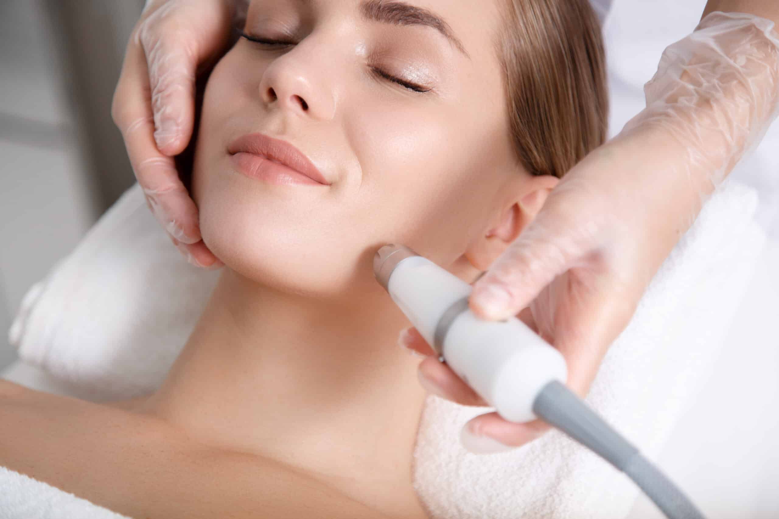 A woman receiving a facial treatment in a spa. A professional in gloves is using a handheld device on her jawline, while the woman reclines comfortably with her eyes closed, wrapped in a towel.