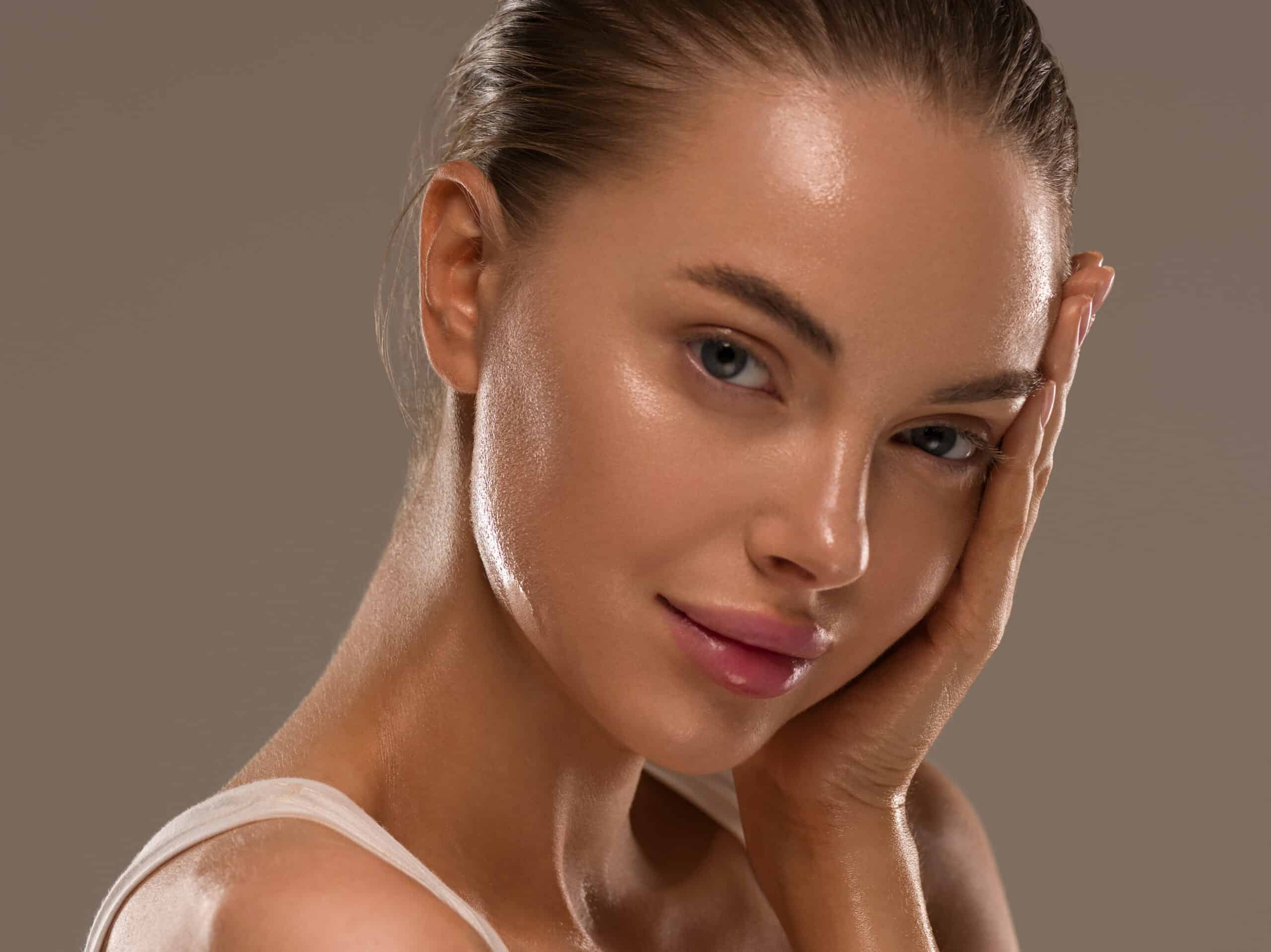 A close-up portrait of a woman with fair skin, softly illuminated. She is wearing minimal makeup, showcasing a natural look. One hand rests on her cheek, and she is gazing slightly towards the camera with a relaxed expression.