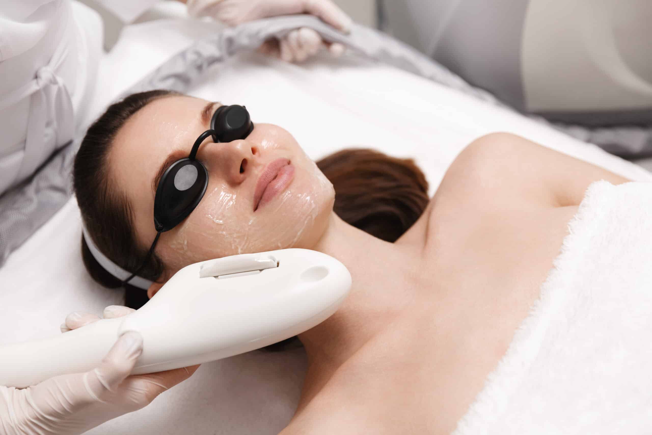 A woman receiving a facial treatment in a spa setting. She wears protective goggles and has a gel-like substance applied to her face. A technician holds a device near her face, indicating a skincare procedure.
