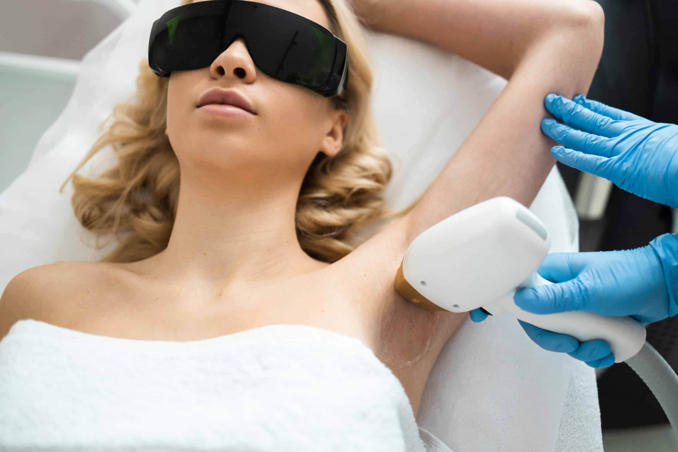 A woman with blonde hair wearing protective glasses lies on a treatment bed as a technician applies a laser hair removal device to her underarm. The woman is wrapped in a towel, and the technician is wearing blue gloves.