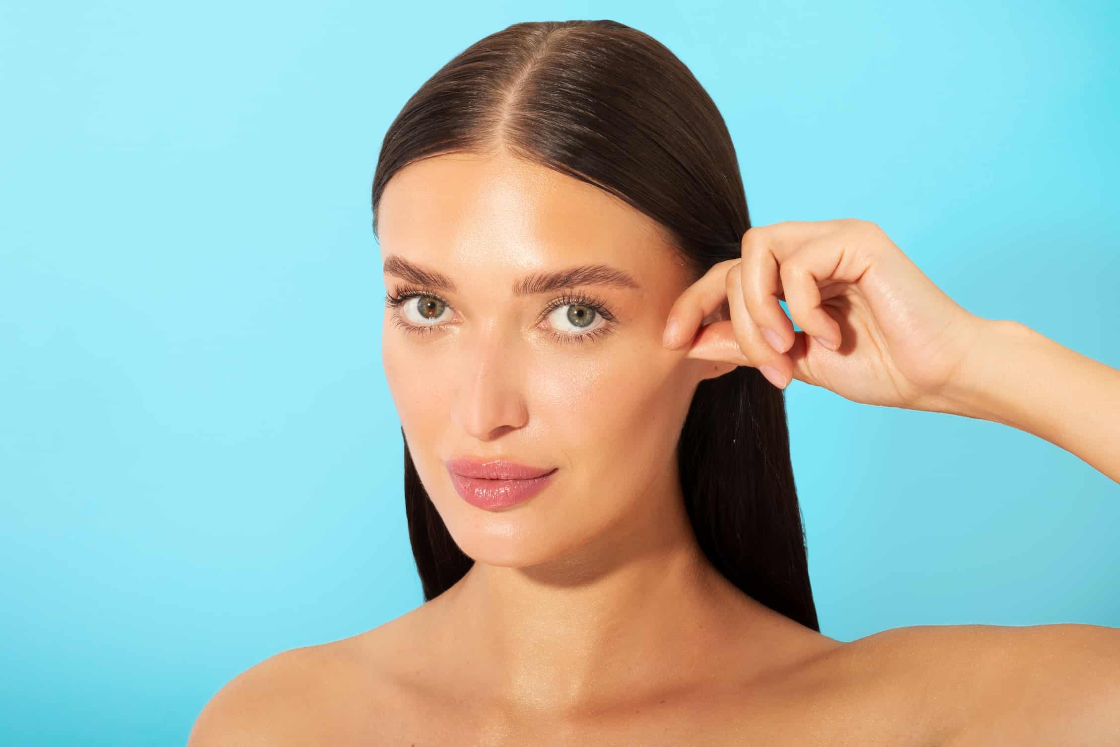 A woman with long, straight dark hair poses against a blue background. She lightly touches her cheek with her right index finger, looking directly at the camera with a neutral expression. Her skin appears smooth and radiant.