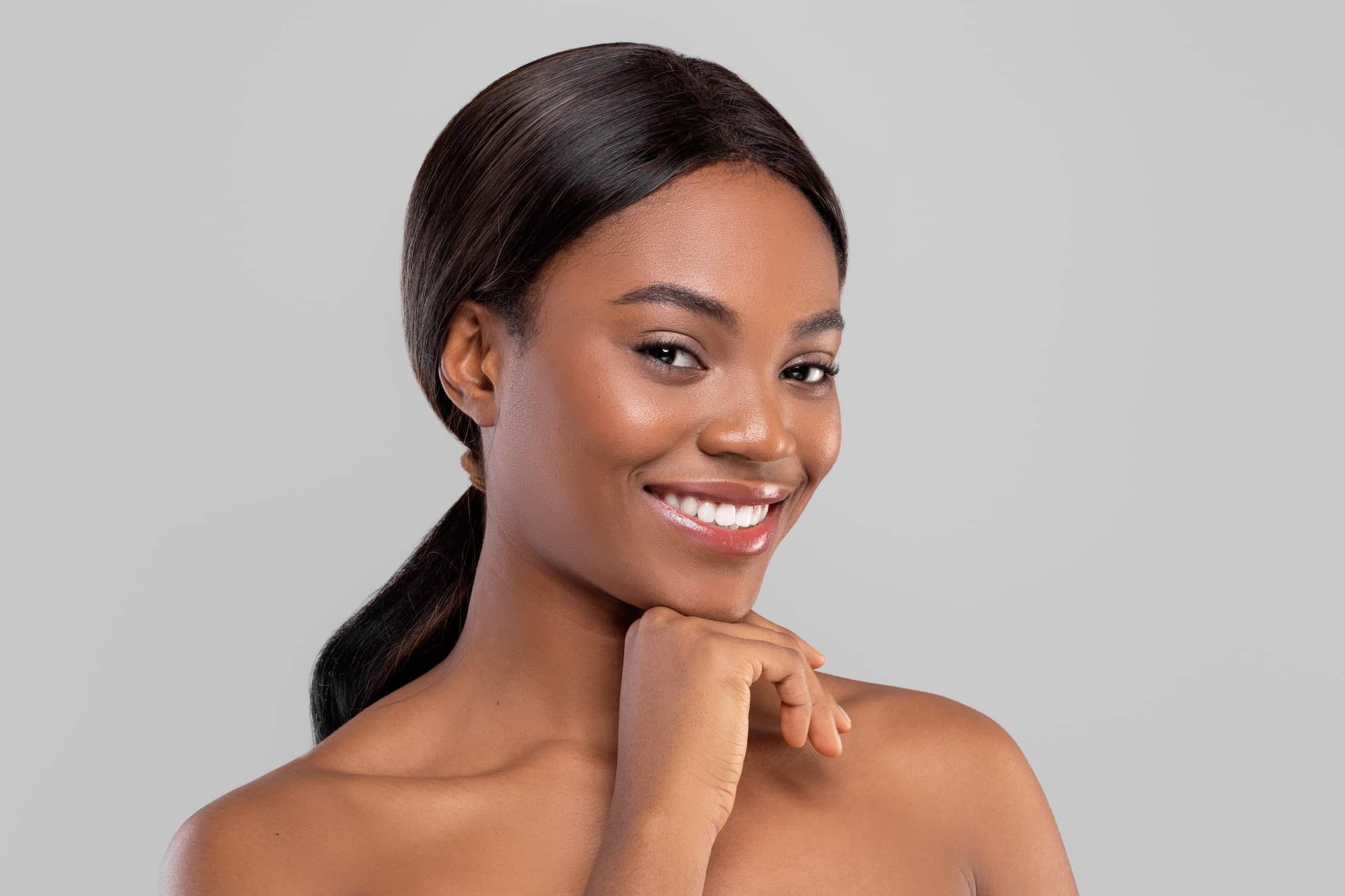 A woman with dark hair styled in a ponytail, facing forward and smiling. She has a natural makeup look and is resting her chin on her hand against a light gray background.