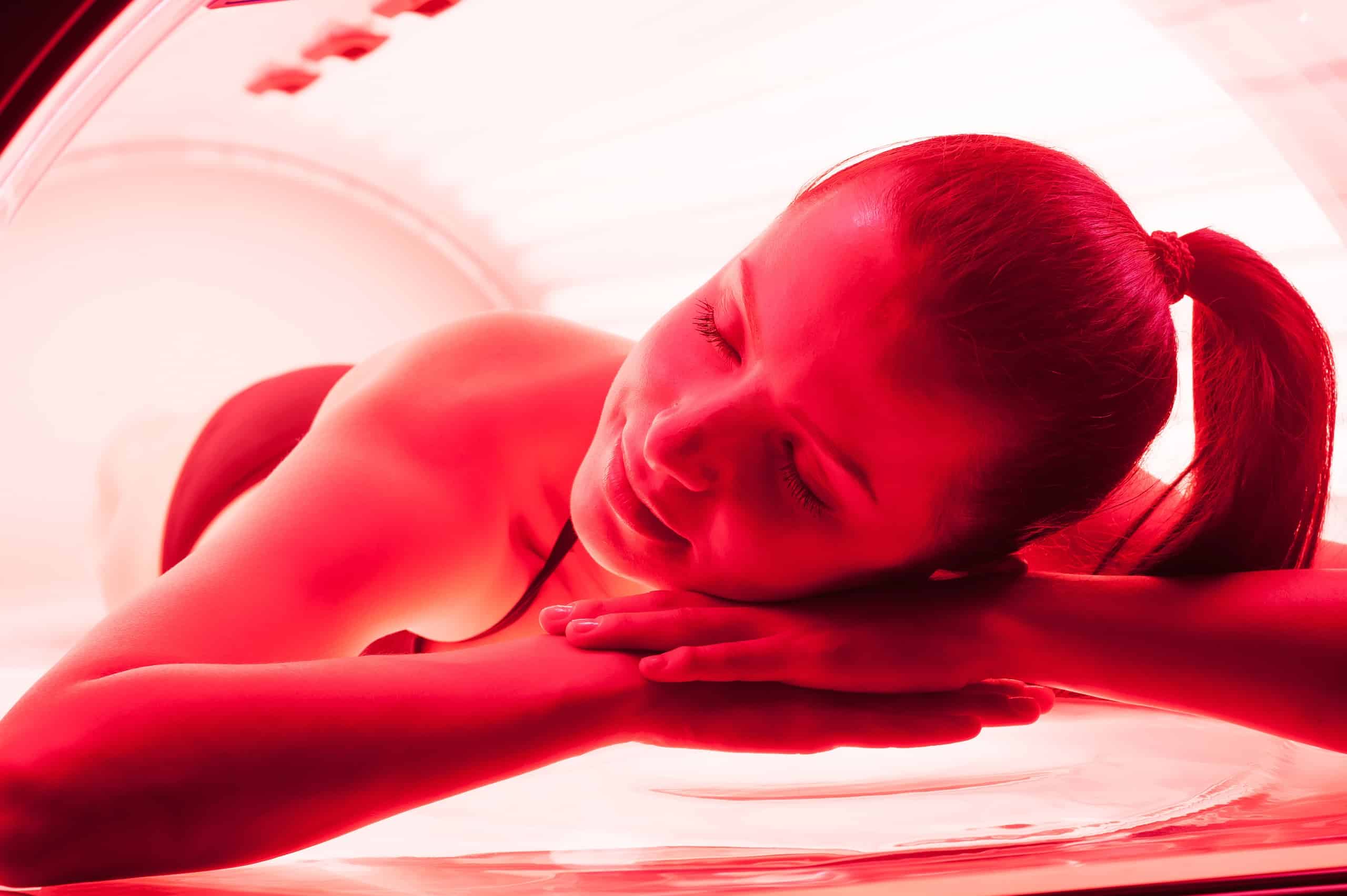 A woman lying inside a tanning bed with red lights, relaxing with her arms folded under her chin. She has a ponytail and appears to be in a serene state, with the interior of the tanning bed softly illuminated around her.