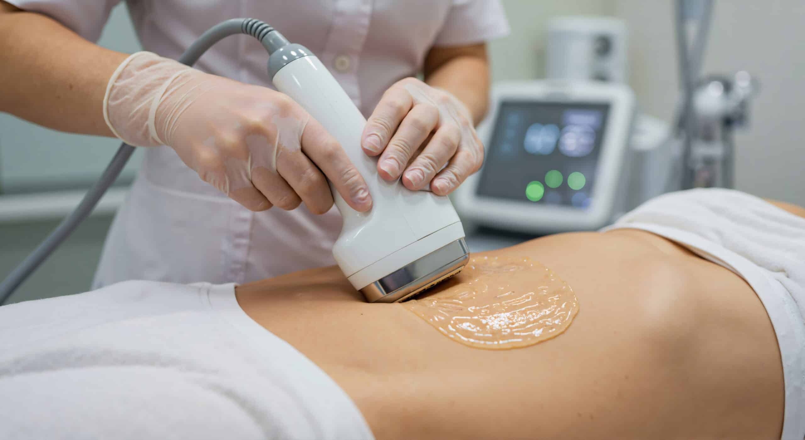 A medical professional using a handheld device for a treatment on a client's abdomen covered with a gel. The practitioner is wearing gloves, and a monitor is visible in the background, indicating a clinical environment.