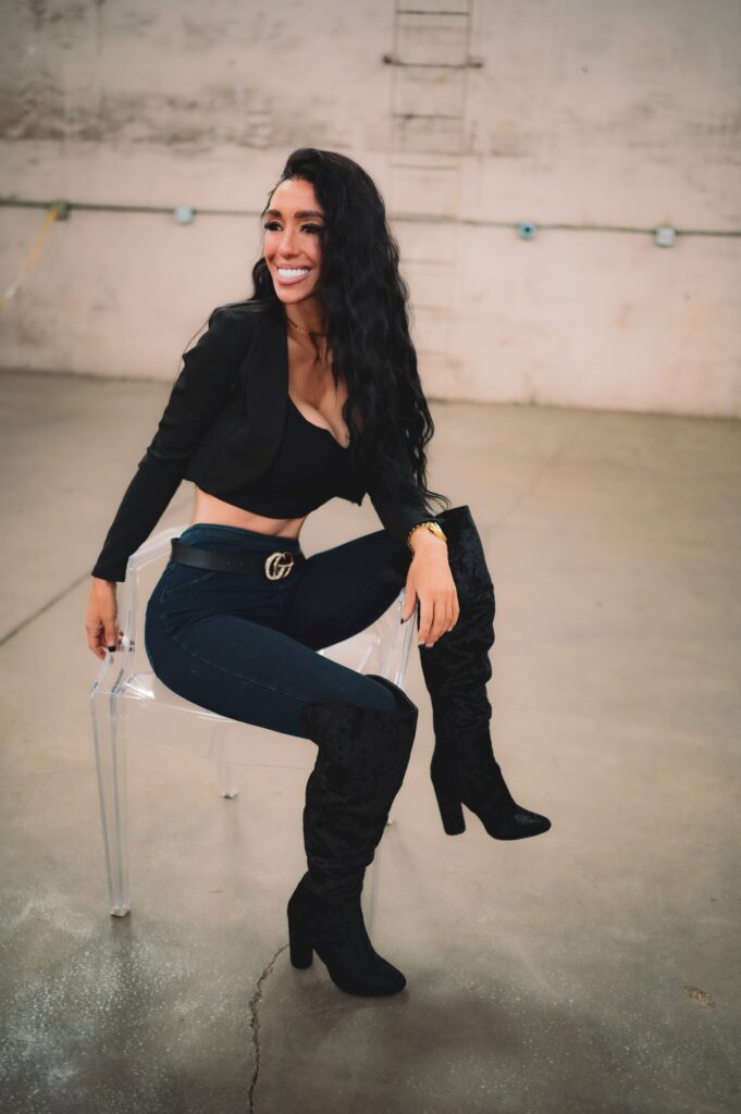 A woman with long black hair smiles while sitting on a clear chair in a minimalistic space. She wears a black cropped jacket, a black top, and dark blue jeans, complemented by tall black boots.