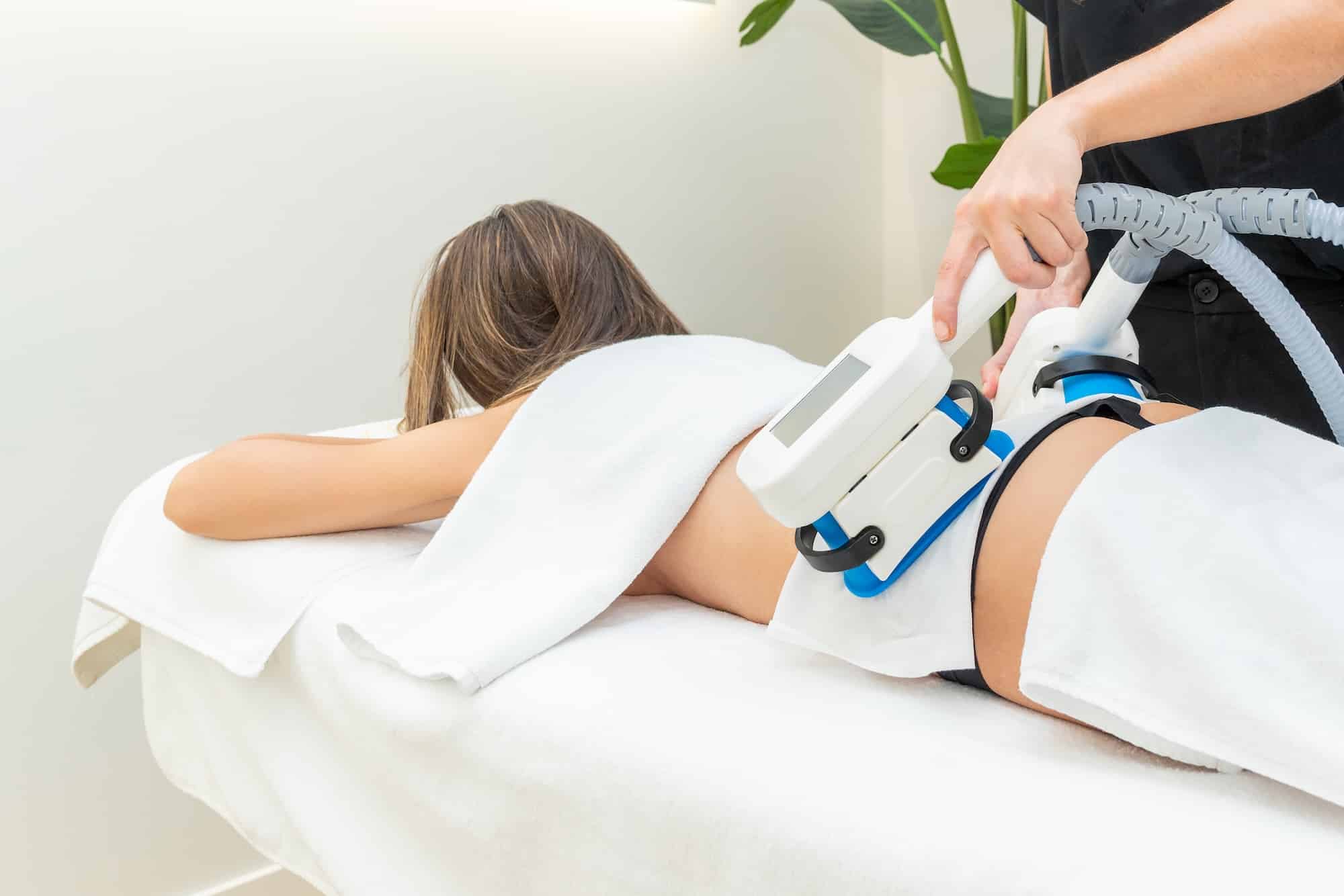 A woman lies on a treatment table, covered with towels, while a technician uses a handheld device on her back. The room has plants in the background, creating a spa-like atmosphere. The focus is on the procedure being performed.