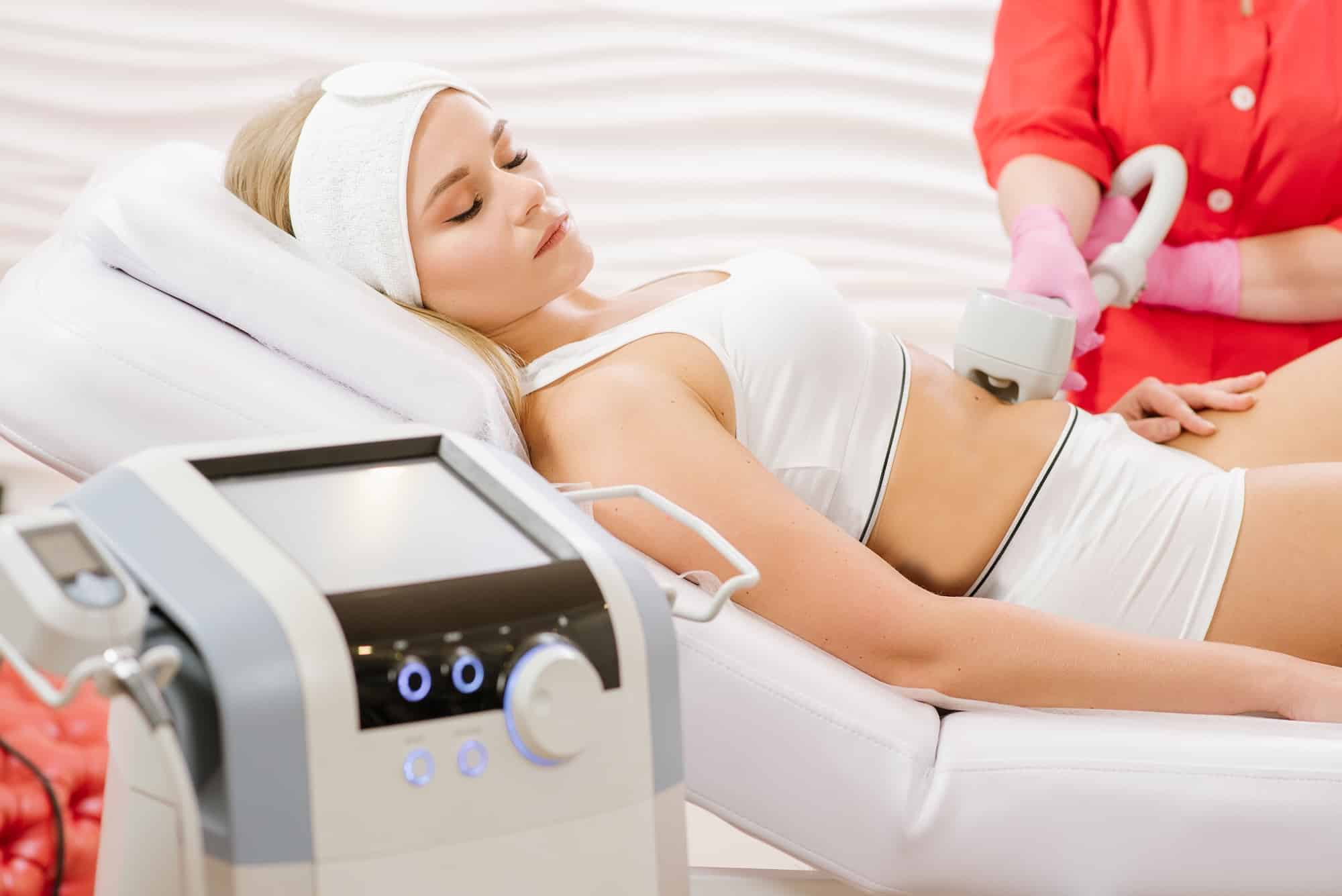A woman lying on a treatment bed wearing a white top and headband, receiving a cosmetic procedure on her abdomen. A technician in gloves operates a device next to her. A modern machine with control panel is visible in the foreground.