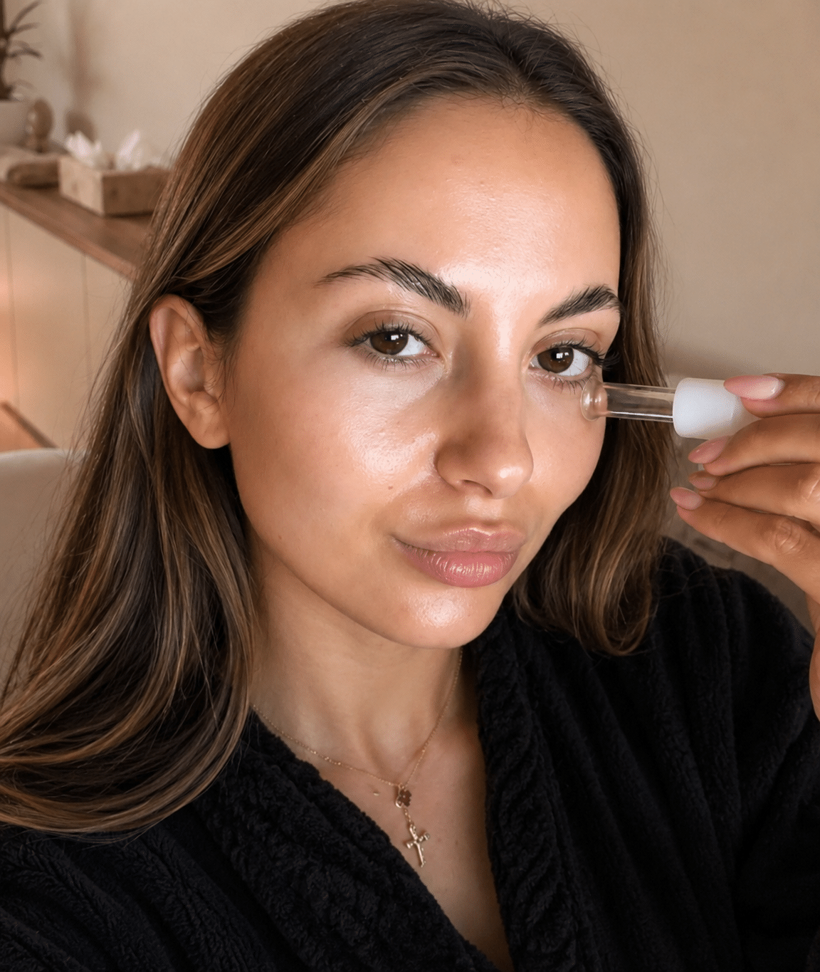A woman with long, brown hair holds a dropper bottle close to her face, applying a clear serum under her eye. She is wearing a black robe, and the background includes a light-colored wall and a wooden surface with decorative items.