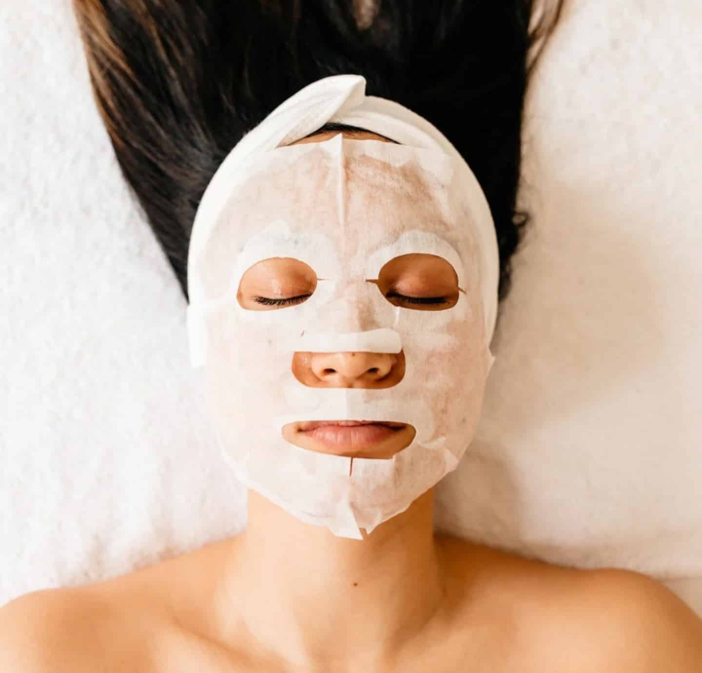A woman lying down with a facial mask covering her face, featuring cut-outs for her eyes and mouth. She has long hair and a headband, and the background is a light-colored surface.