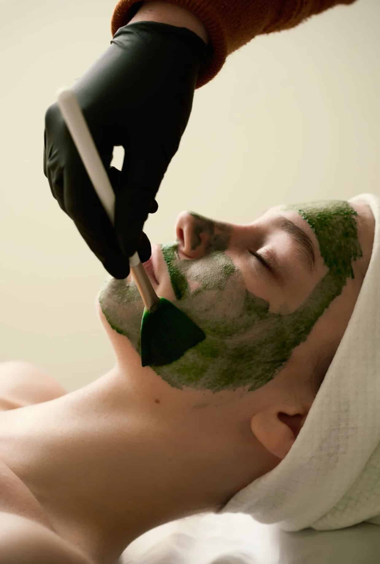 A person lying on a treatment table with a green facial mask applied to their face. A gloved hand is using a brush to apply the mask, while the individual wears a white towel around their head.