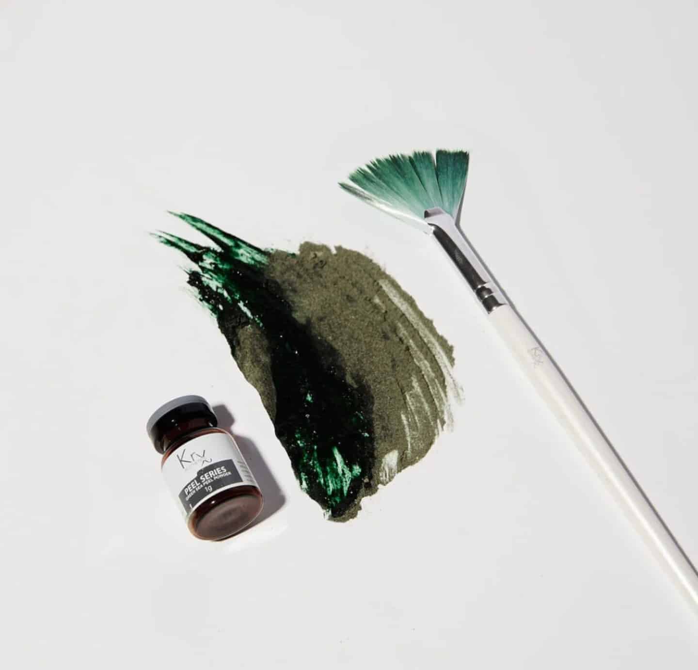 A small jar of dark green paint is placed near a brush with a teal handle, alongside a thick, textured stroke of dark green paint on white paper. The brush shows remnants of the same paint color.