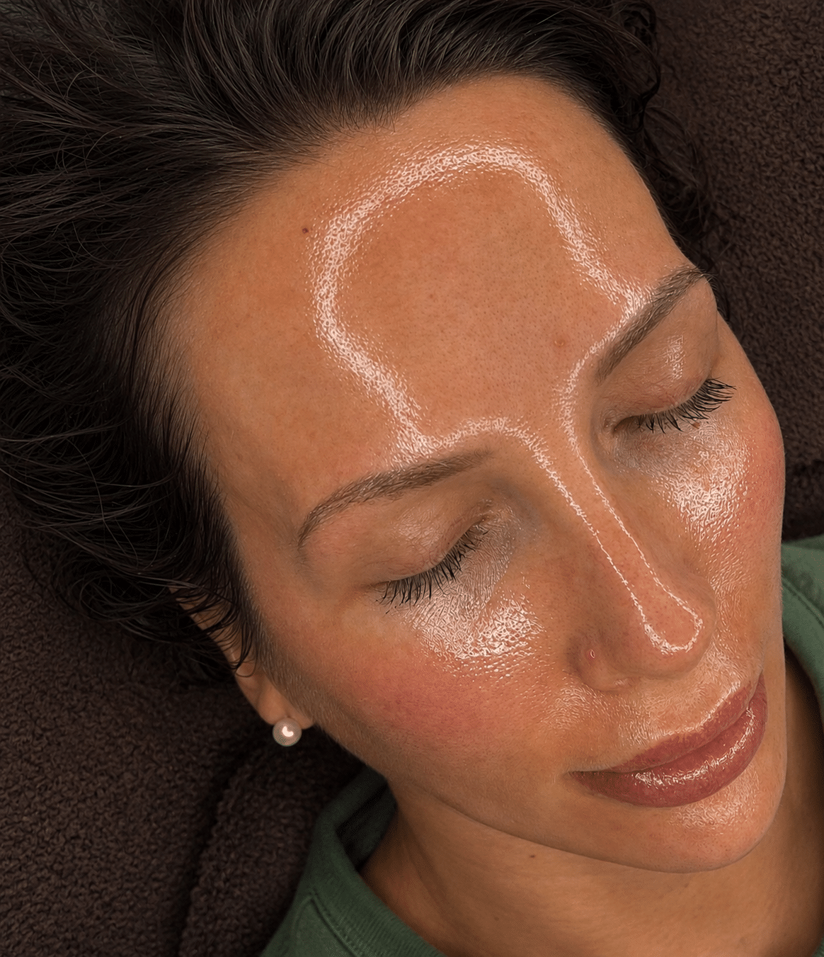 A woman lies with her eyes closed, showing her face covered in a transparent gel with a smooth finish. She has short dark hair and wears a green top, with pearl earrings visible.