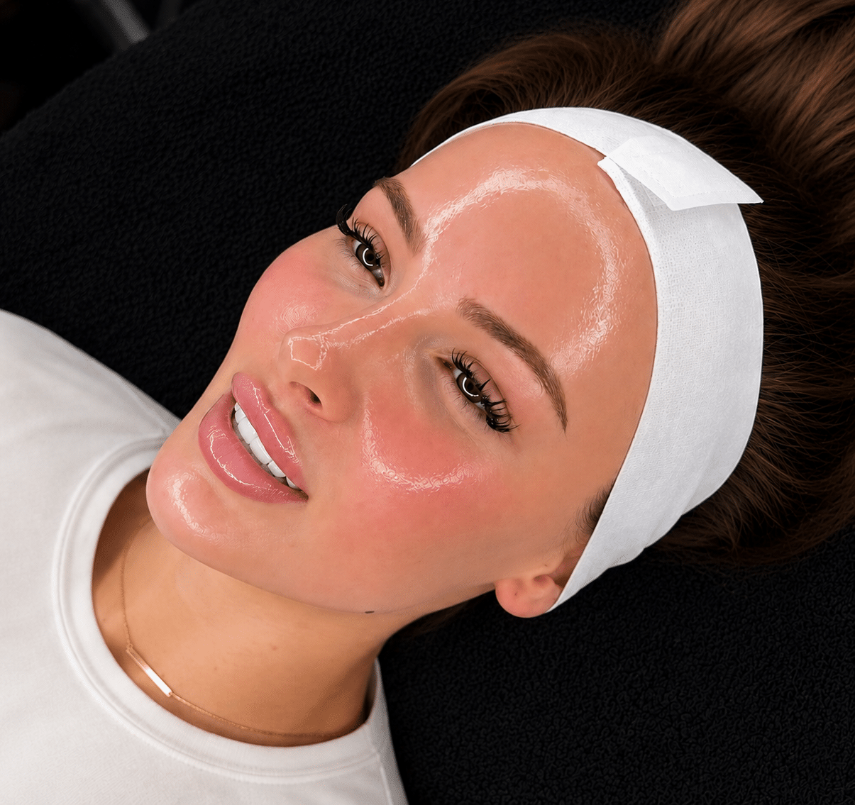 A woman lying on a treatment table with a white headband, skin covered in a clear facial mask. She is wearing a light-colored top and is positioned against a dark backdrop.