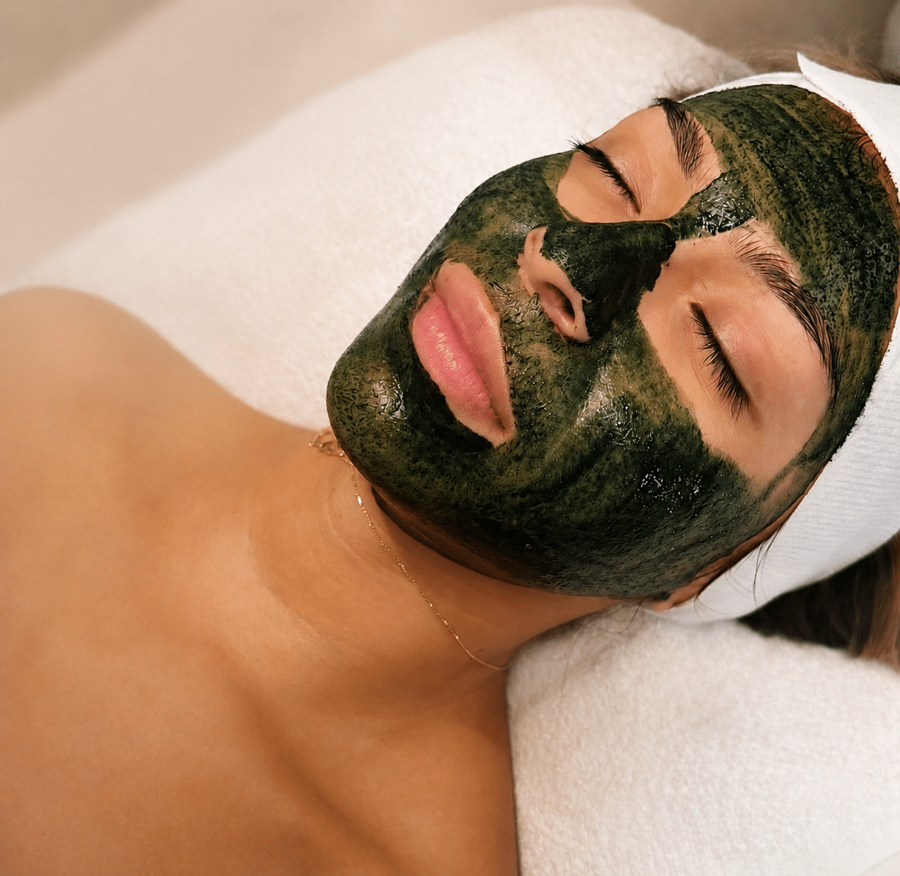 A woman with a green facial mask lies on a treatment bed, wearing a white headband. Her eyes are closed, and her upper body is covered with a soft blanket, while a light background suggests a relaxed spa environment.