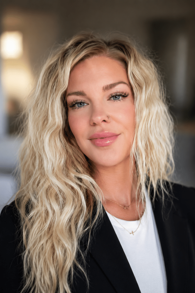 A woman with long, wavy blonde hair wearing a black blazer over a white top stands facing the camera. The background is softly blurred, indicating a well-lit indoor setting.