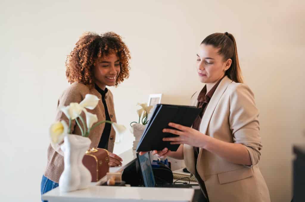 A woman with curly hair and a tan sweater stands beside another woman in a blazer, discussing items at a counter. The counter has decorative flowers and a tablet, with a muted background.