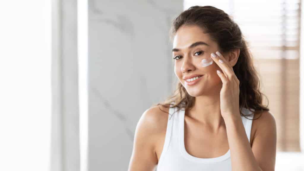 A woman in a white tank top applies cream to her cheek in a bright bathroom with a window and blinds in the background.