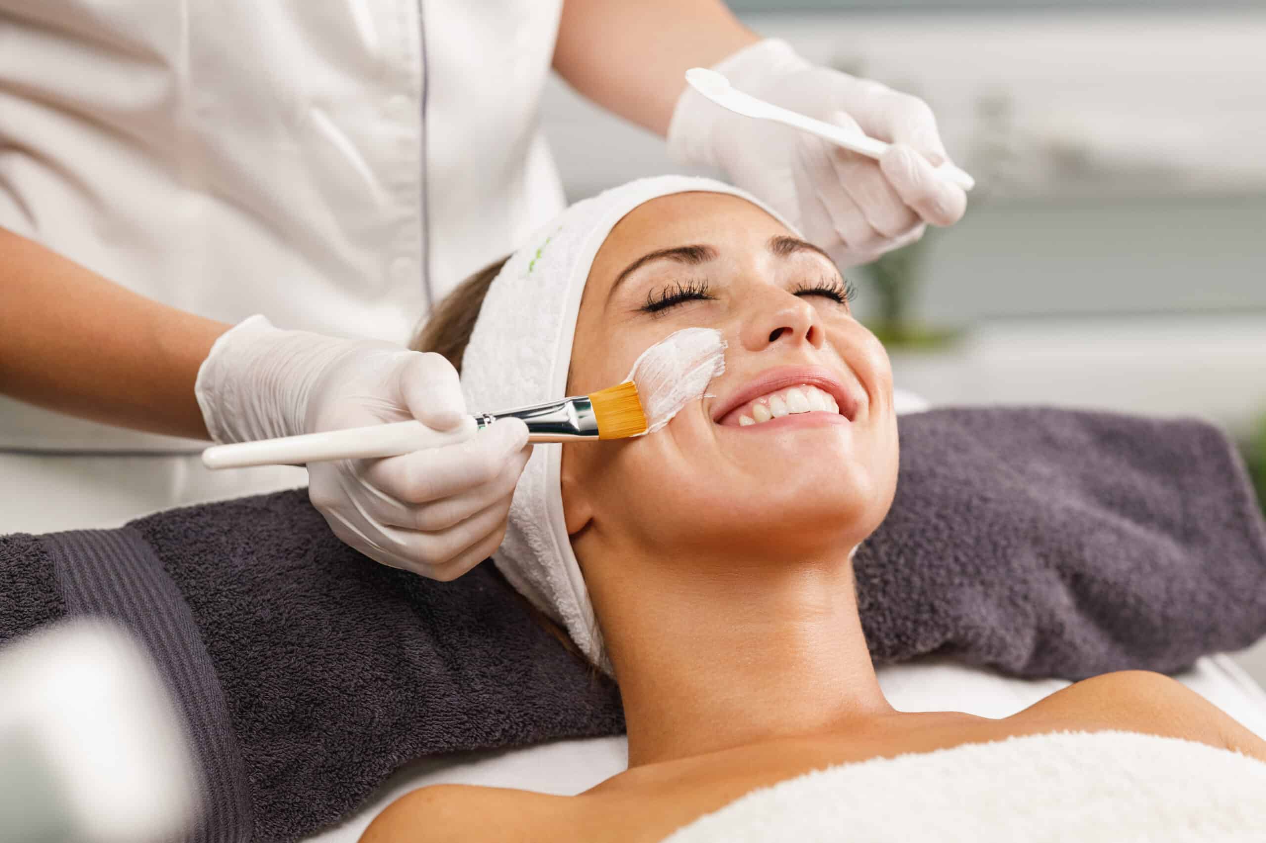 A woman receives a facial treatment with a brush on her cheek while lying on a spa bed with a white headband and towel.