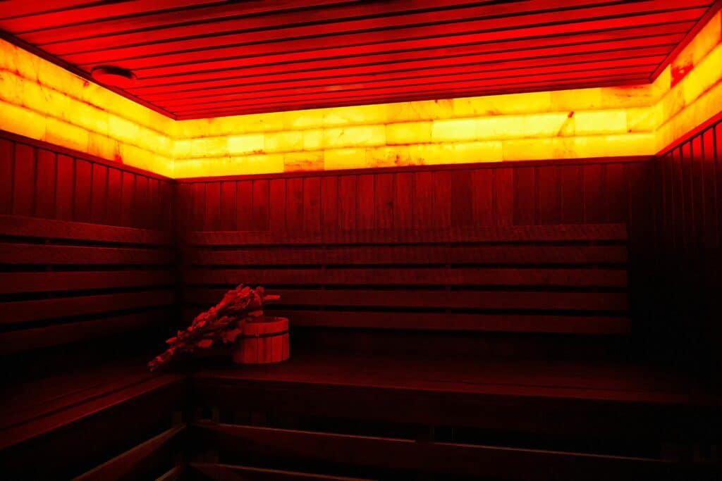 A red-lit sauna with wooden benches and slatted walls, featuring a small bucket with leafy branches on the floor.